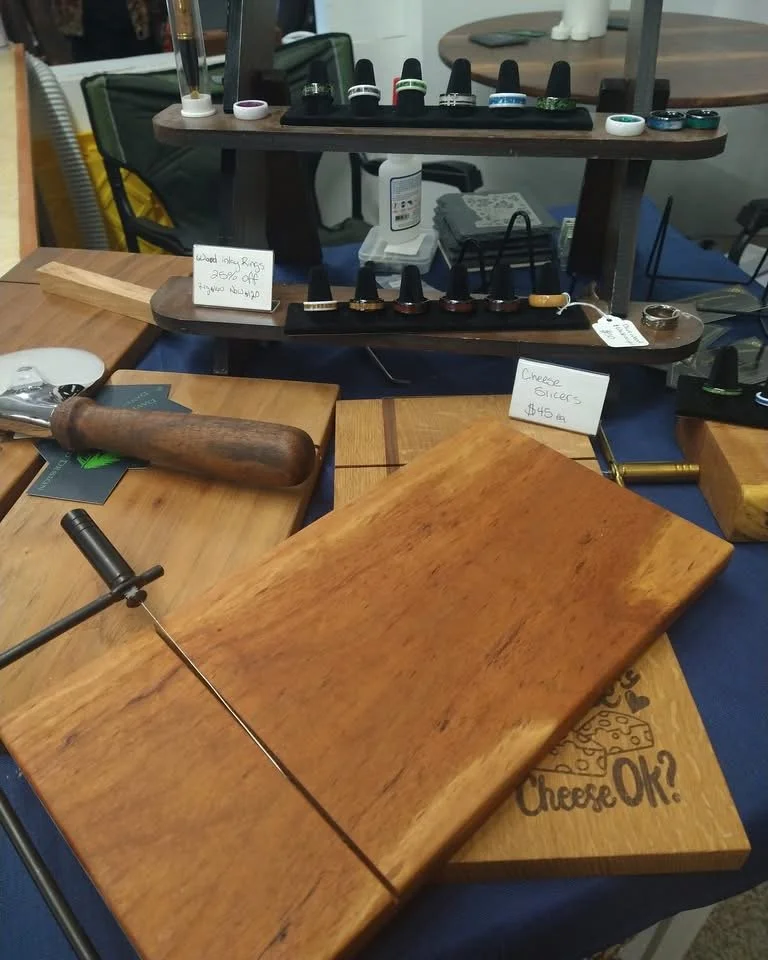 Display of nail polishes and rings on a wooden stand at a market or craft fair, with a leather rolling pin, wooden cutting boards, and a jewelry price tag on a table.