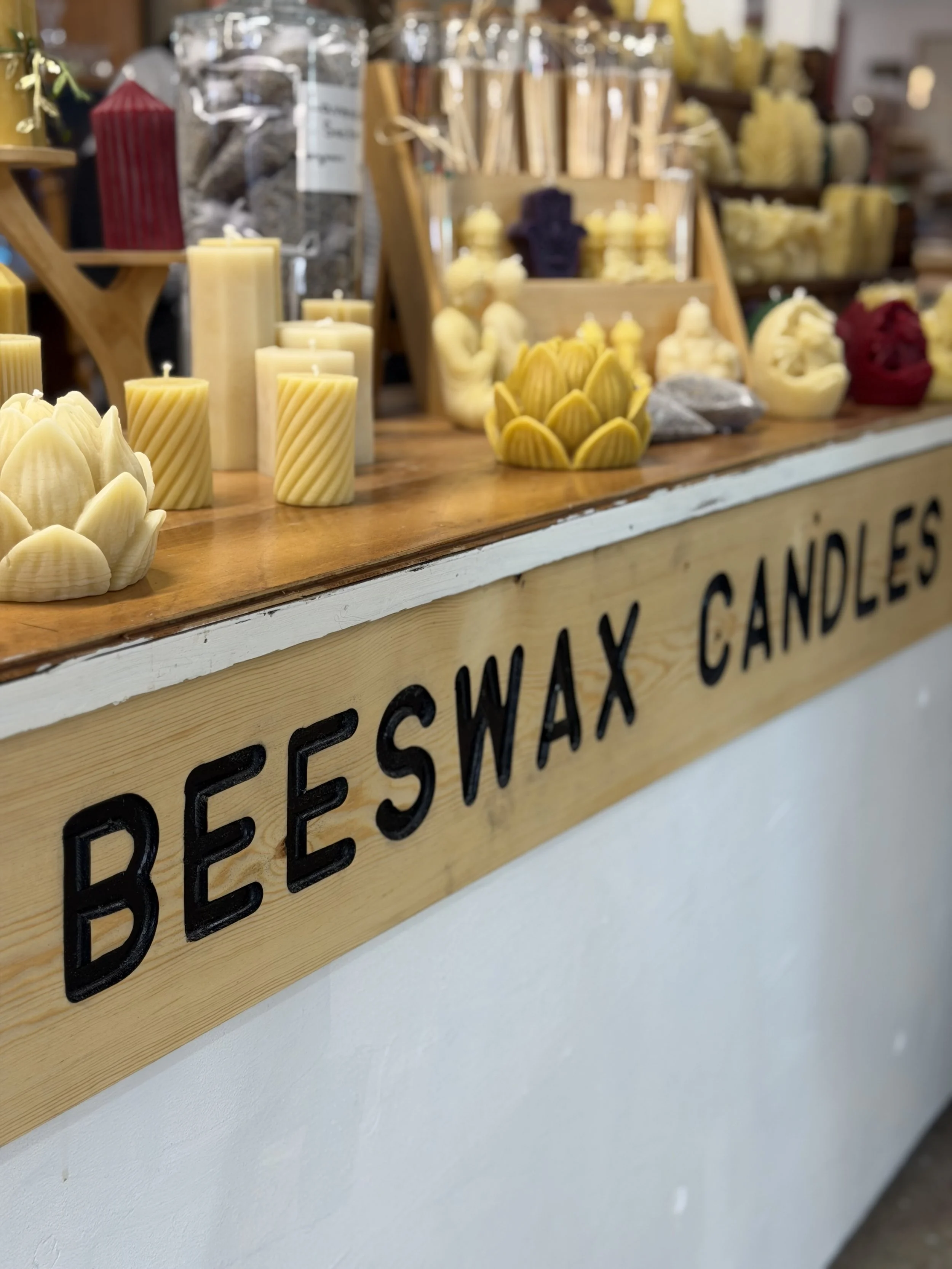 Close-up of a wooden display shelf with beeswax candles in various shapes and sizes for sale, including some in the shape of flowers and others in tall, cylindrical forms.