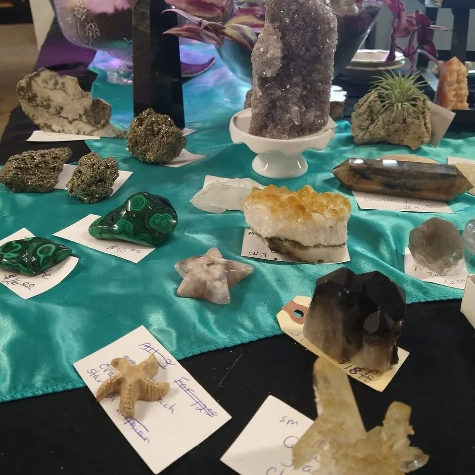 Display of various minerals and crystals on a teal fabric surface, each with handwritten tags indicating their names and prices. Features large and small specimens, including a starfish-shaped mineral, a green malachite stone, and a black and white crystal cluster.