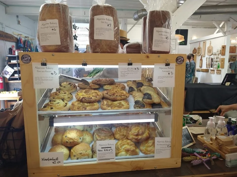 Display case with baked goods at Alperose Bakery, including cinnamon rolls, pastries, and bread, with price tags and packaging above.