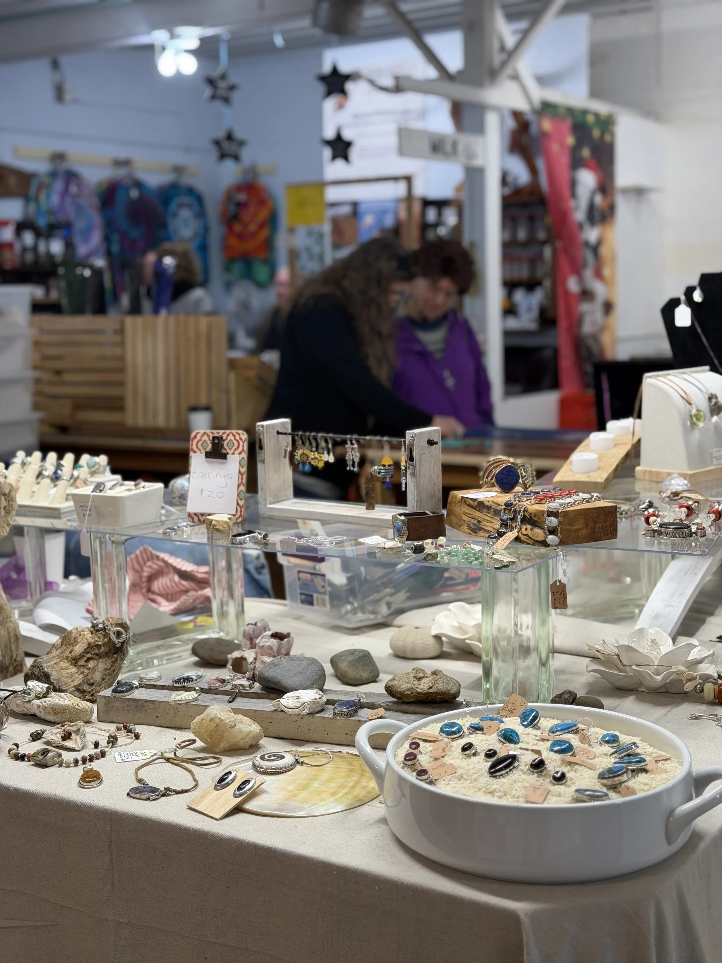 A craft fair display table featuring various jewelry pieces including rings, earrings, and bracelets, with decorative rocks and shells, at an indoor market.