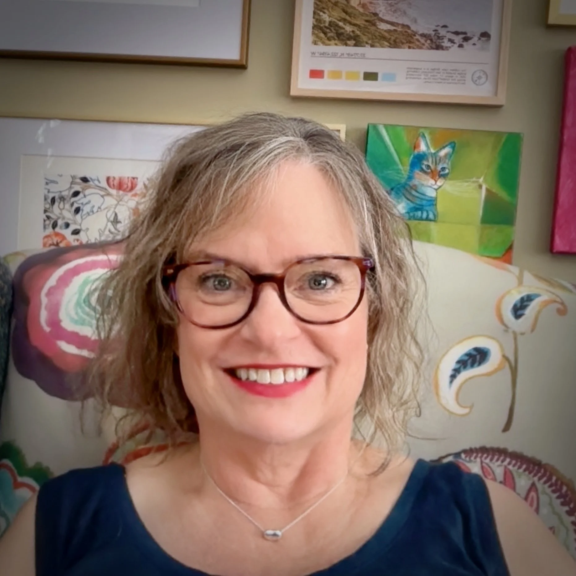 A smiling woman with curly gray hair and glasses, wearing a necklace, sitting in front of colorful artwork and framed posters on a wall.