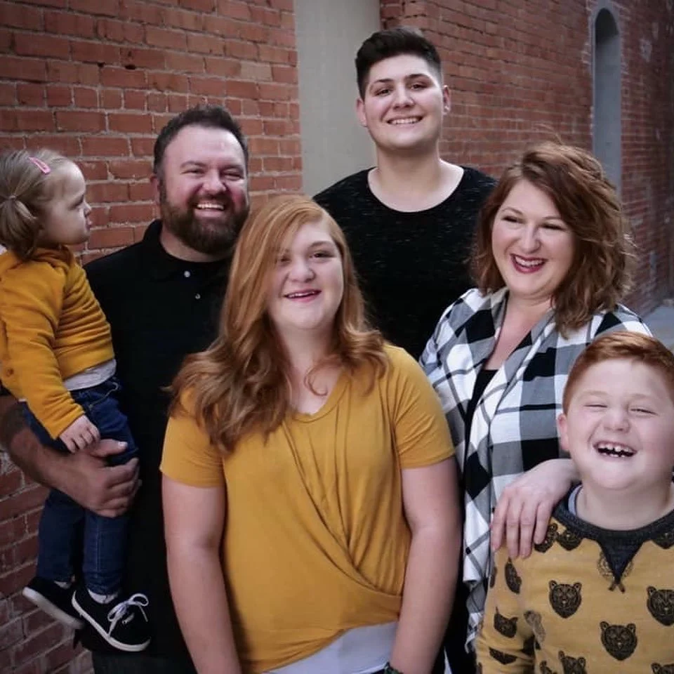 A smiling family of six standing in front of a brick wall, including two children, two women, and two men.