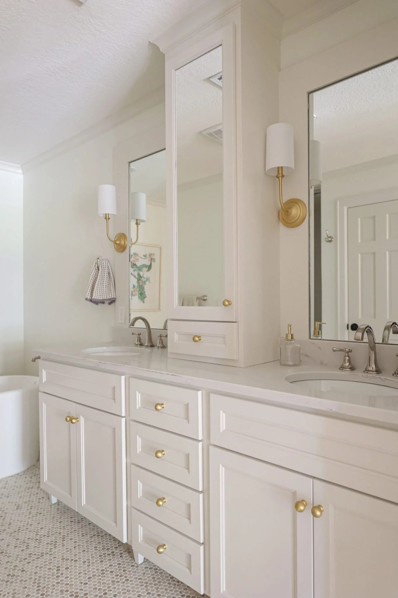 A bathroom vanity with multiple drawers and cabinets, white with gold knobs, a double sink, and two mirrors, with wall-mounted white and gold light fixtures, a framed artwork, and a patterned floor.