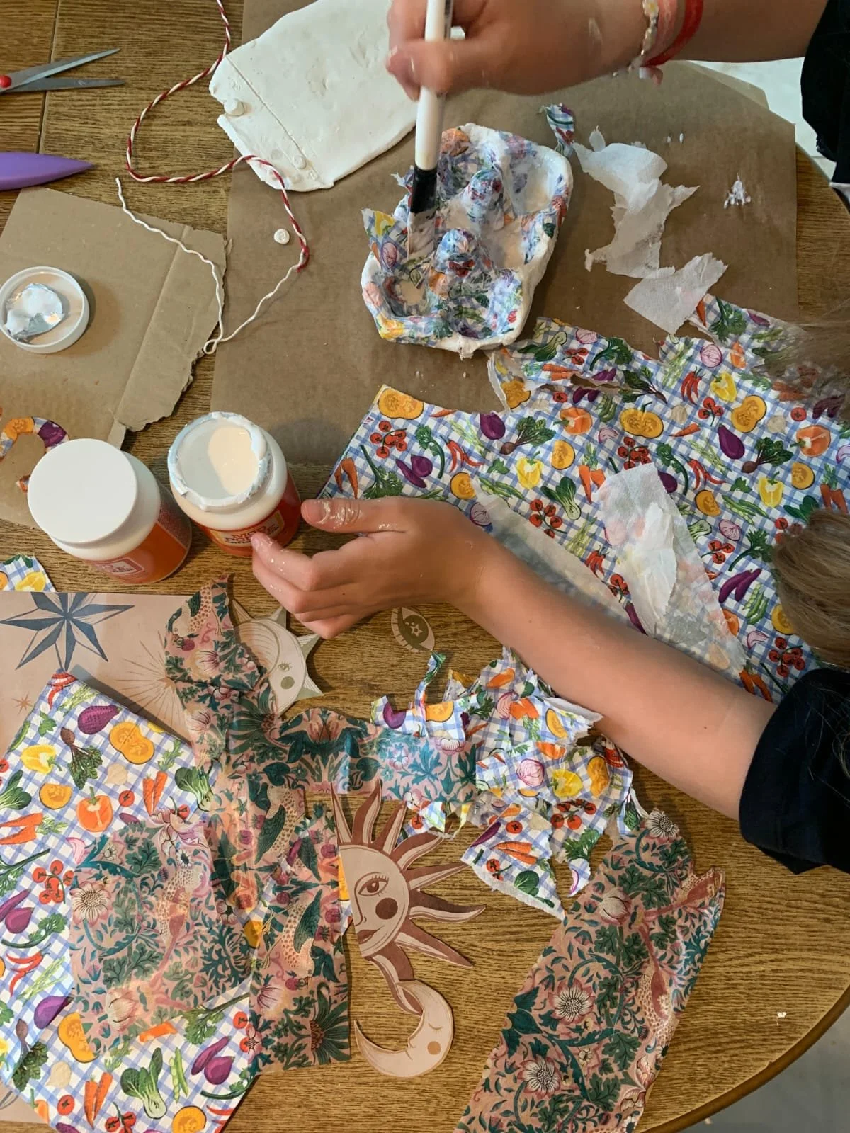 People wrapping a gift with festive, colorful patterned wrapping paper decorated with food items and a sun illustration on a wooden table, surrounded by scissors, tape, and glue.
