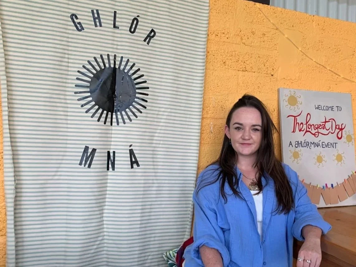 A woman sitting indoors near a wall with a decorative clock and a welcome sign that reads "The Longest Day A GHLÓRMÁNÉ EVENT."