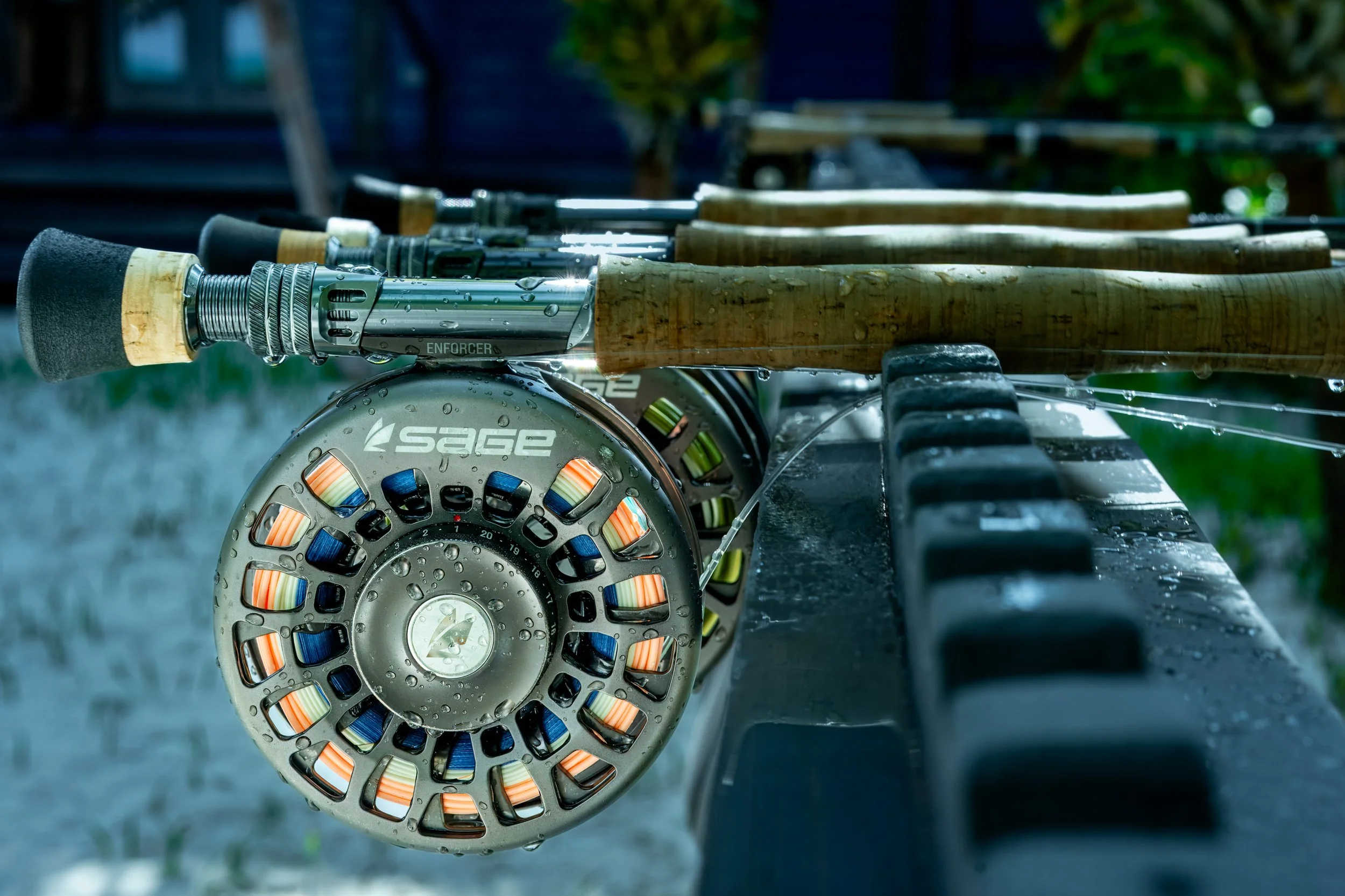 Close-up image of a fishing reel attached to a fishing rod, with water droplets on the reel, placed on a black rack outdoors, with a blurred background of trees.