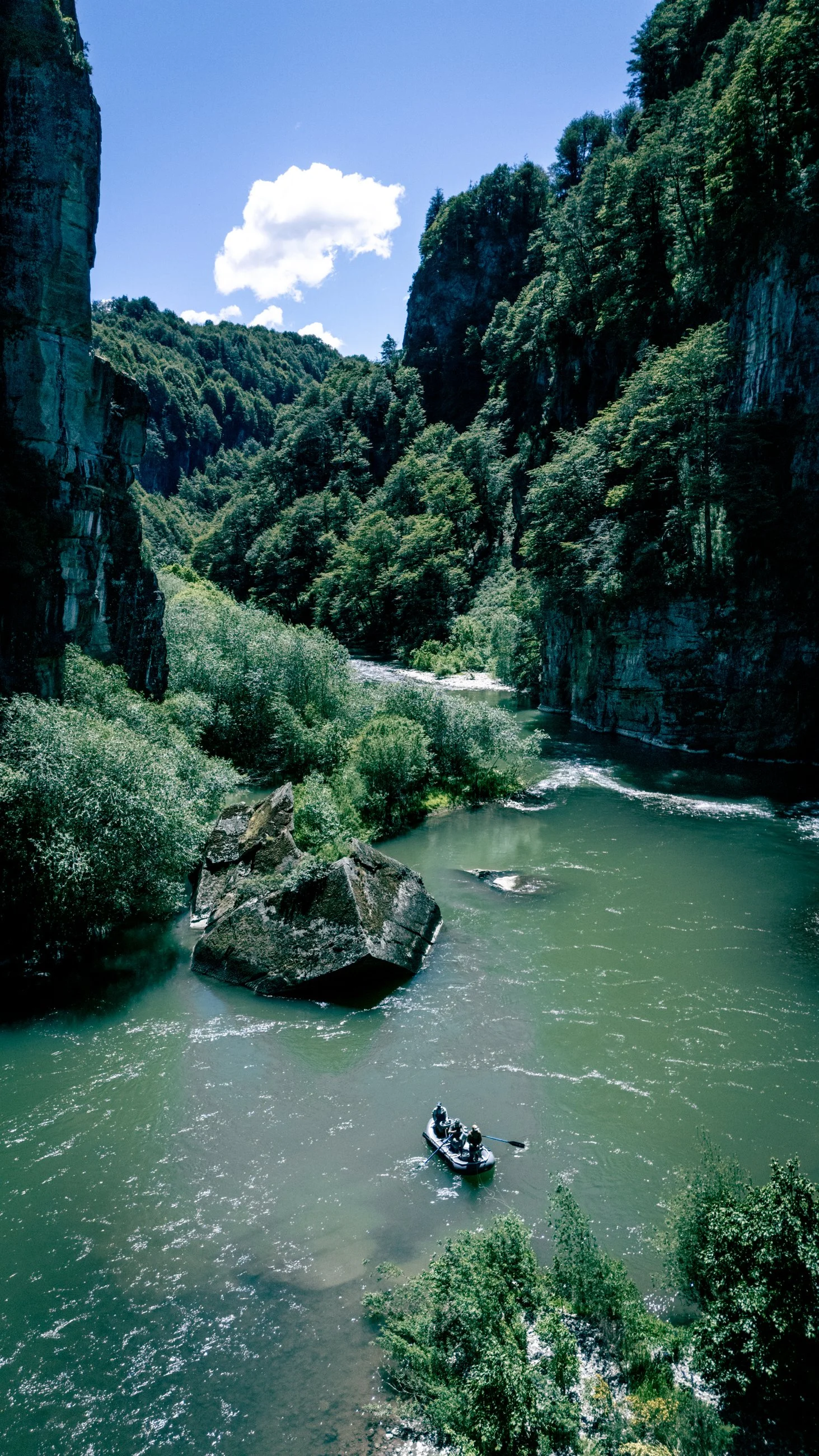 A boat with people on a river flowing through a deep canyon in Chile with steep cliffs and dense green forest at the top, under a bright blue sky with a few clouds.