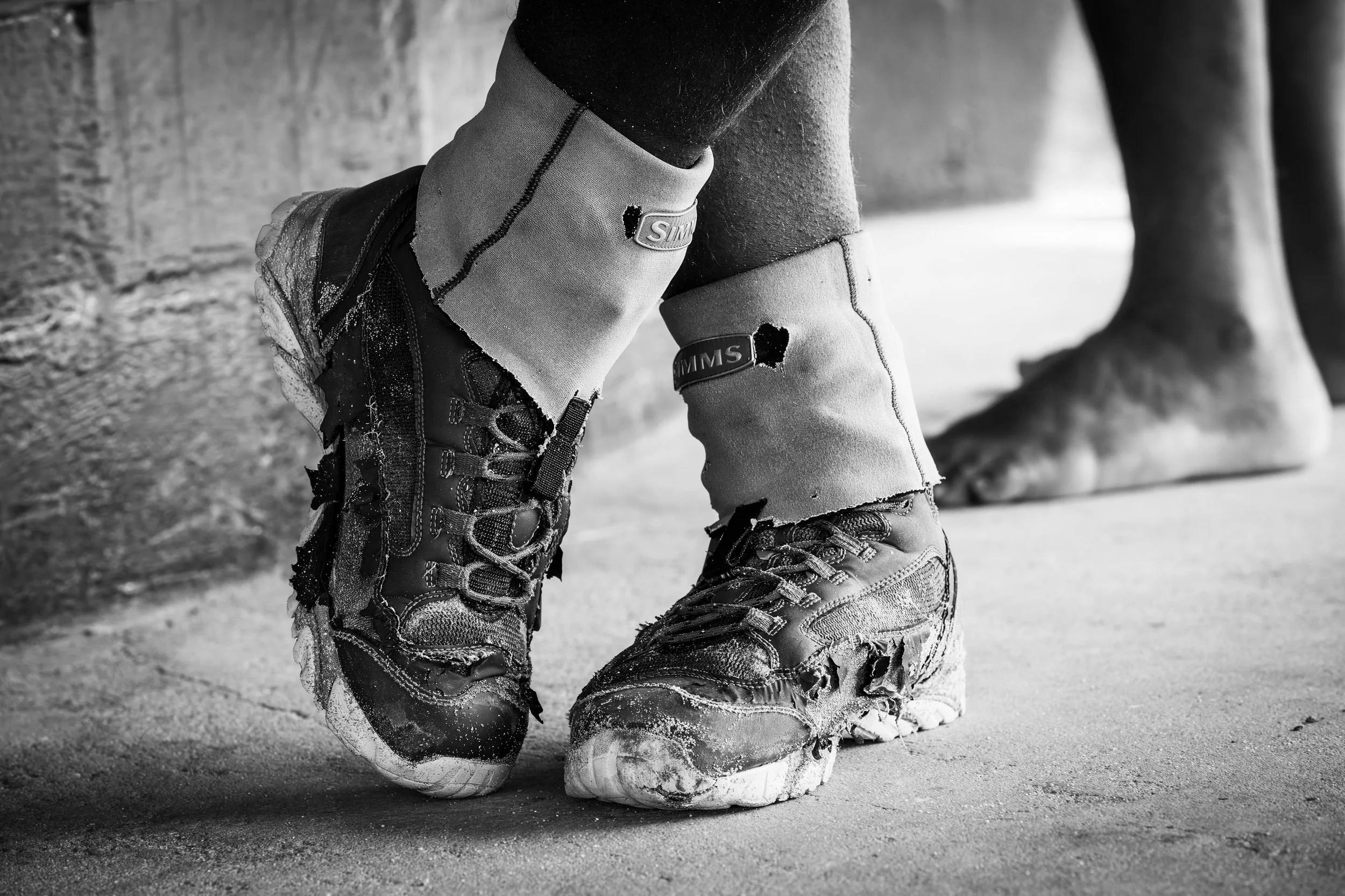 Close-up of a person wearing beat up flats boots and pants, kneeling on snow-covered ground, with snow and a tree trunk in the background.