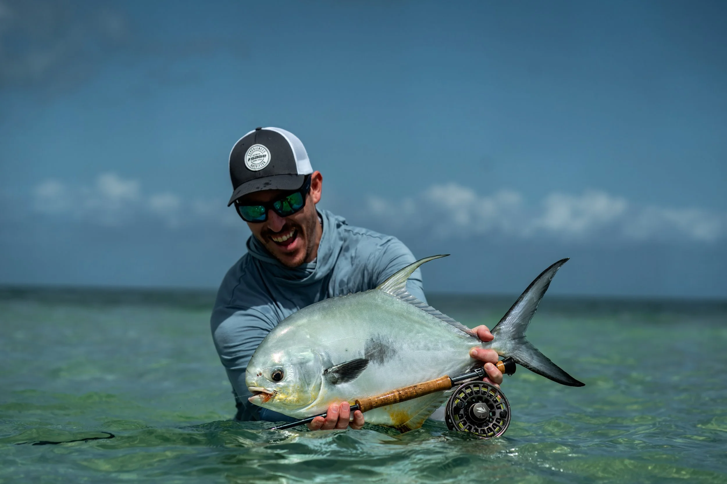A man in sunglasses and a hat holding a permit in Mexico, with a fly fishing rod in hand and a cloudy sky in the background.