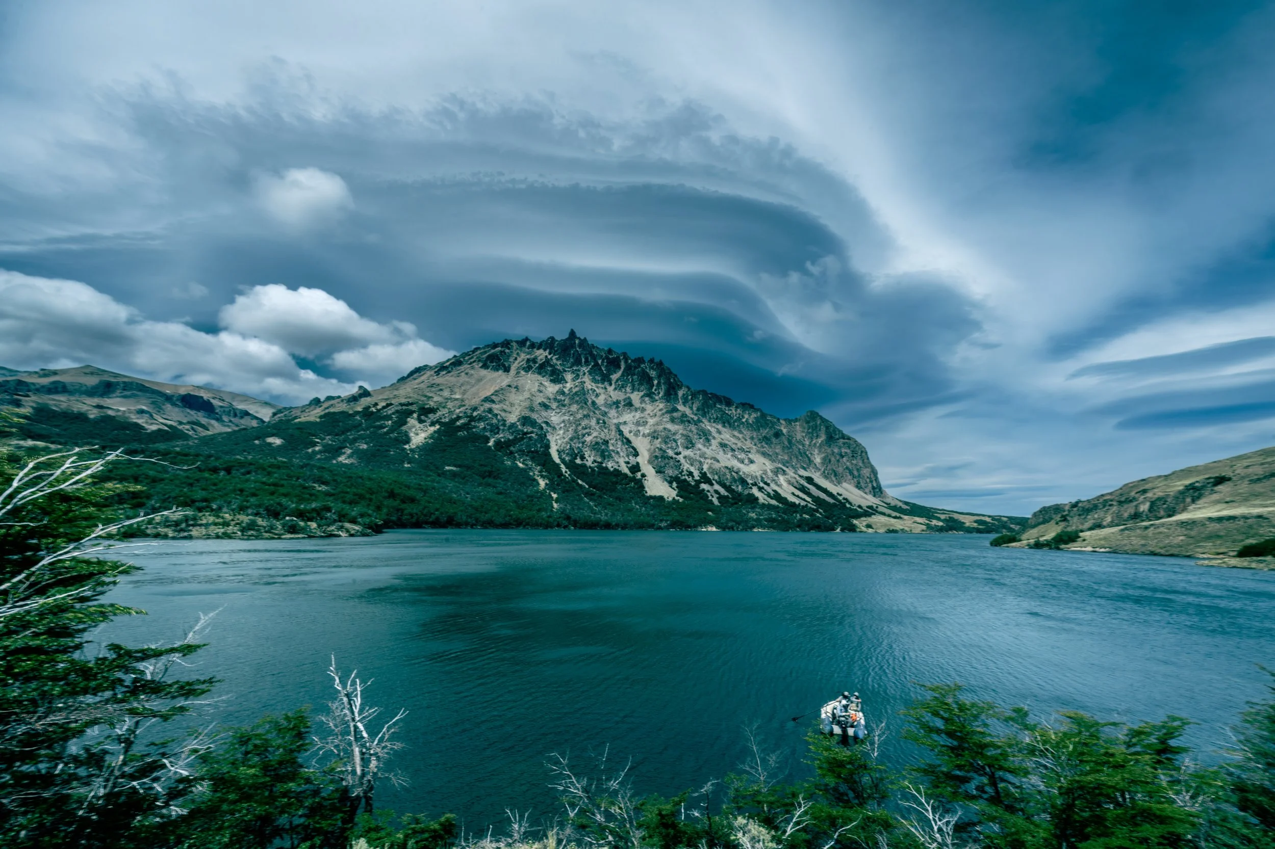 A mountain landscape in Patagonia with a cloudy sky, water in the foreground, and a small boat with people on the water.