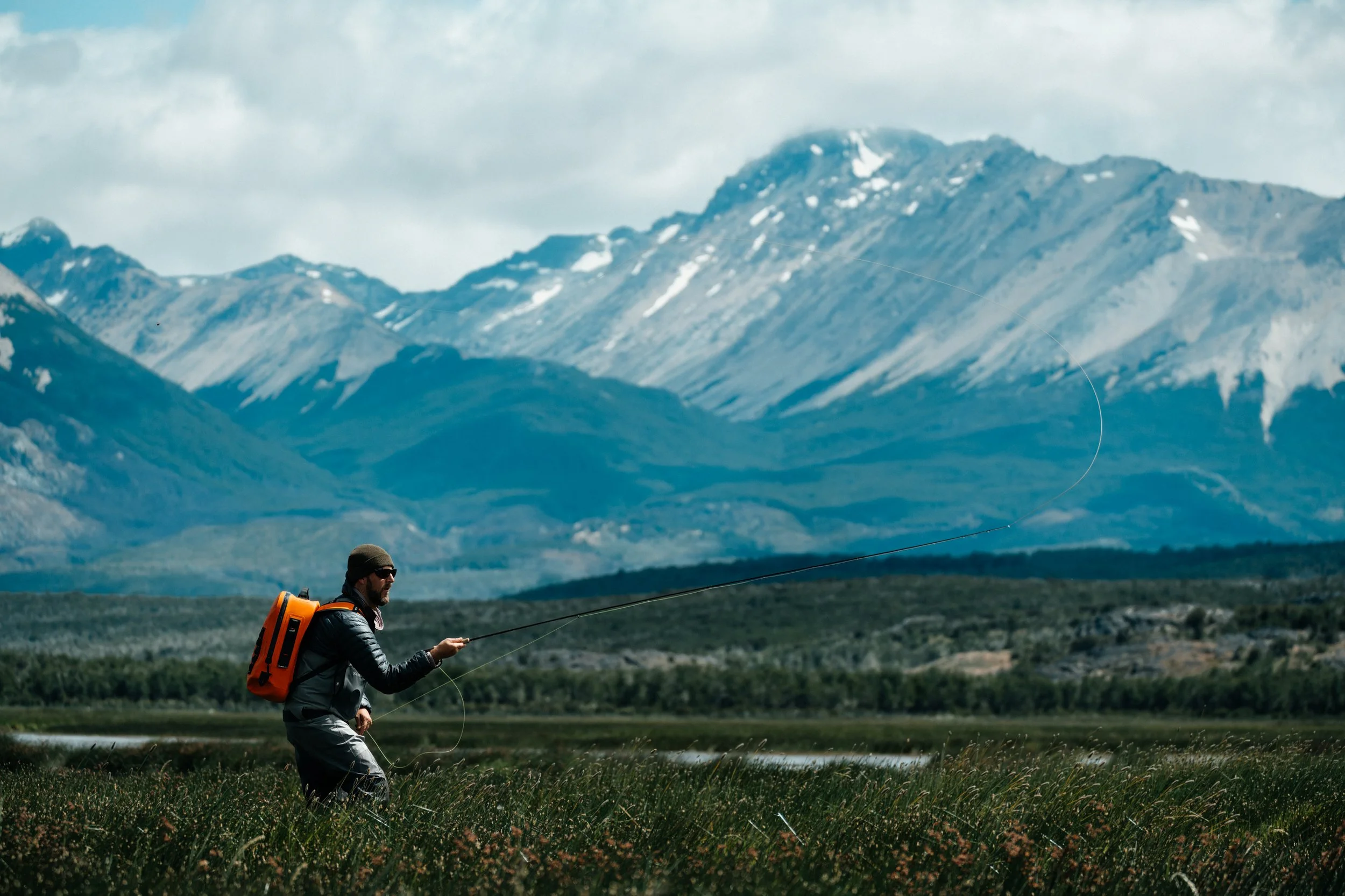 A man fishing in a river with a backdrop of snow-capped mountains and cloudy sky in Patagonia.