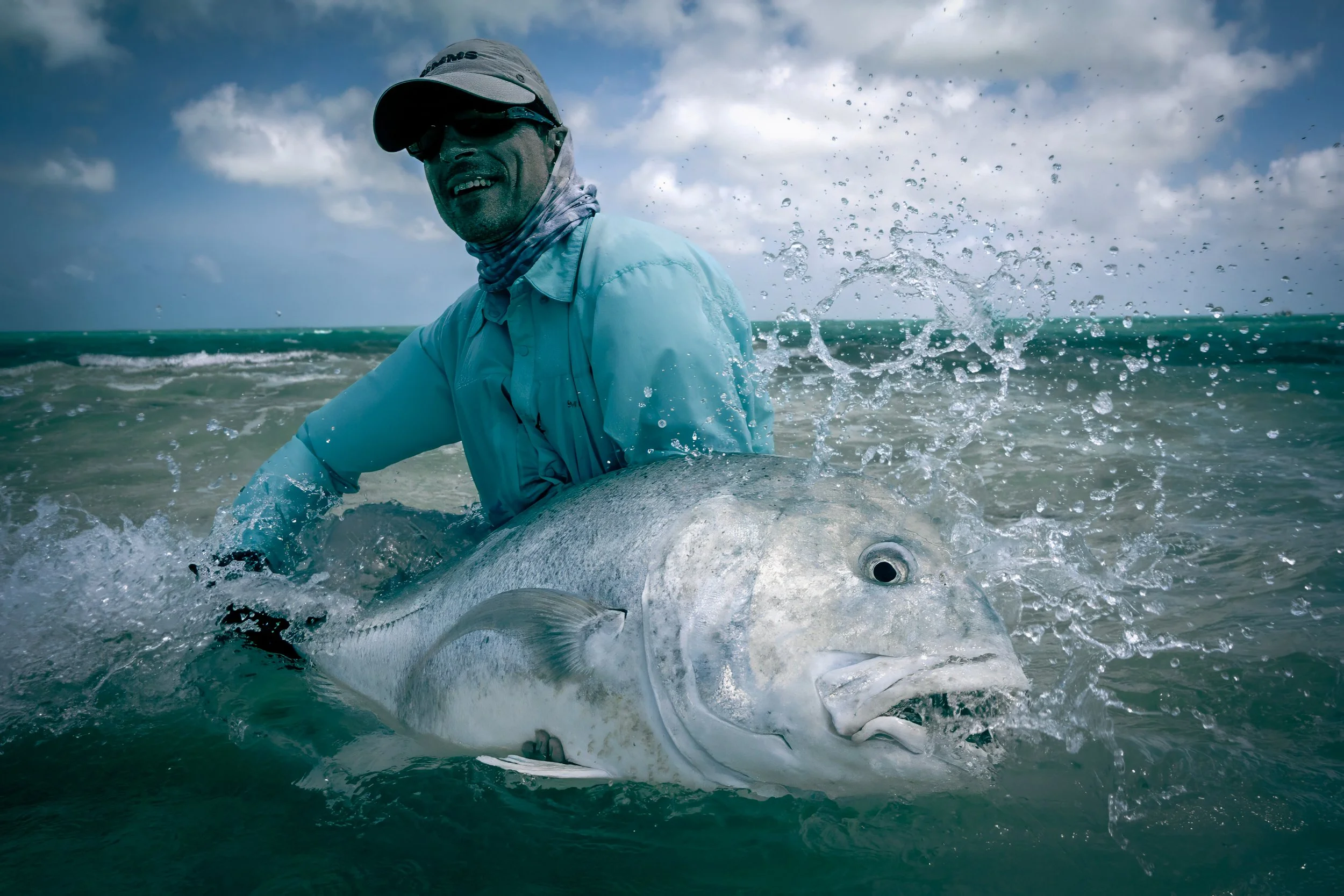A man in sun protective clothing and sunglasses holding a Giant Trevall in Christmas Island, with waves splashing around.