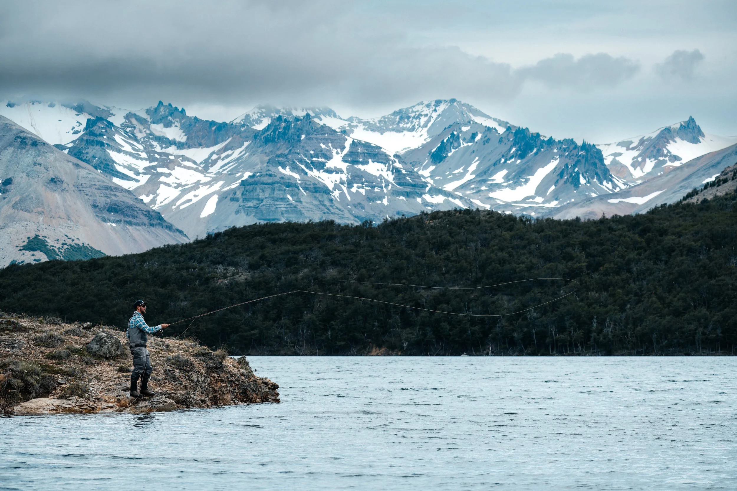 A man fishing by a lake in Patagonia with snow-capped mountains in the background under a cloudy sky.