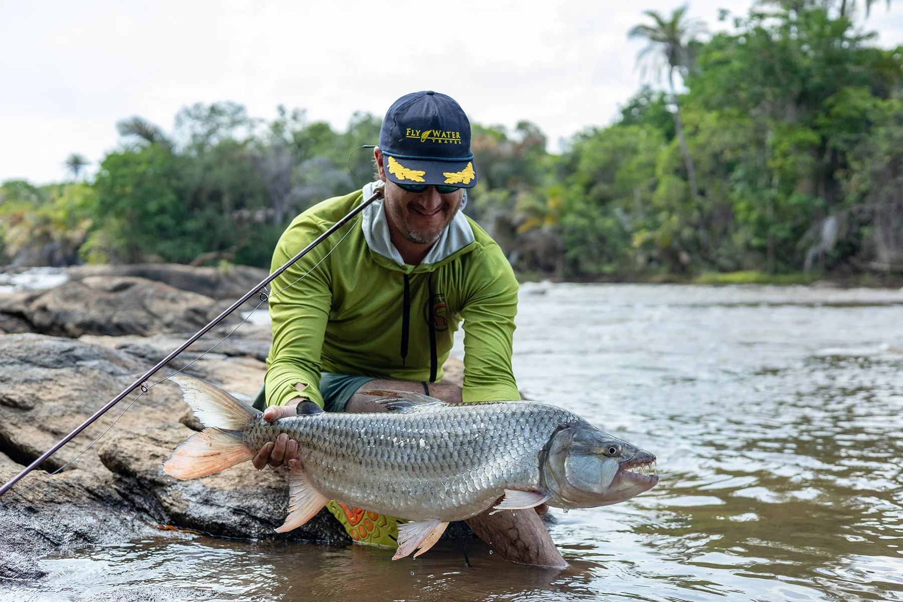 Man in a green jacket and blue cap holding a large fish near a riverbank with trees in the background.