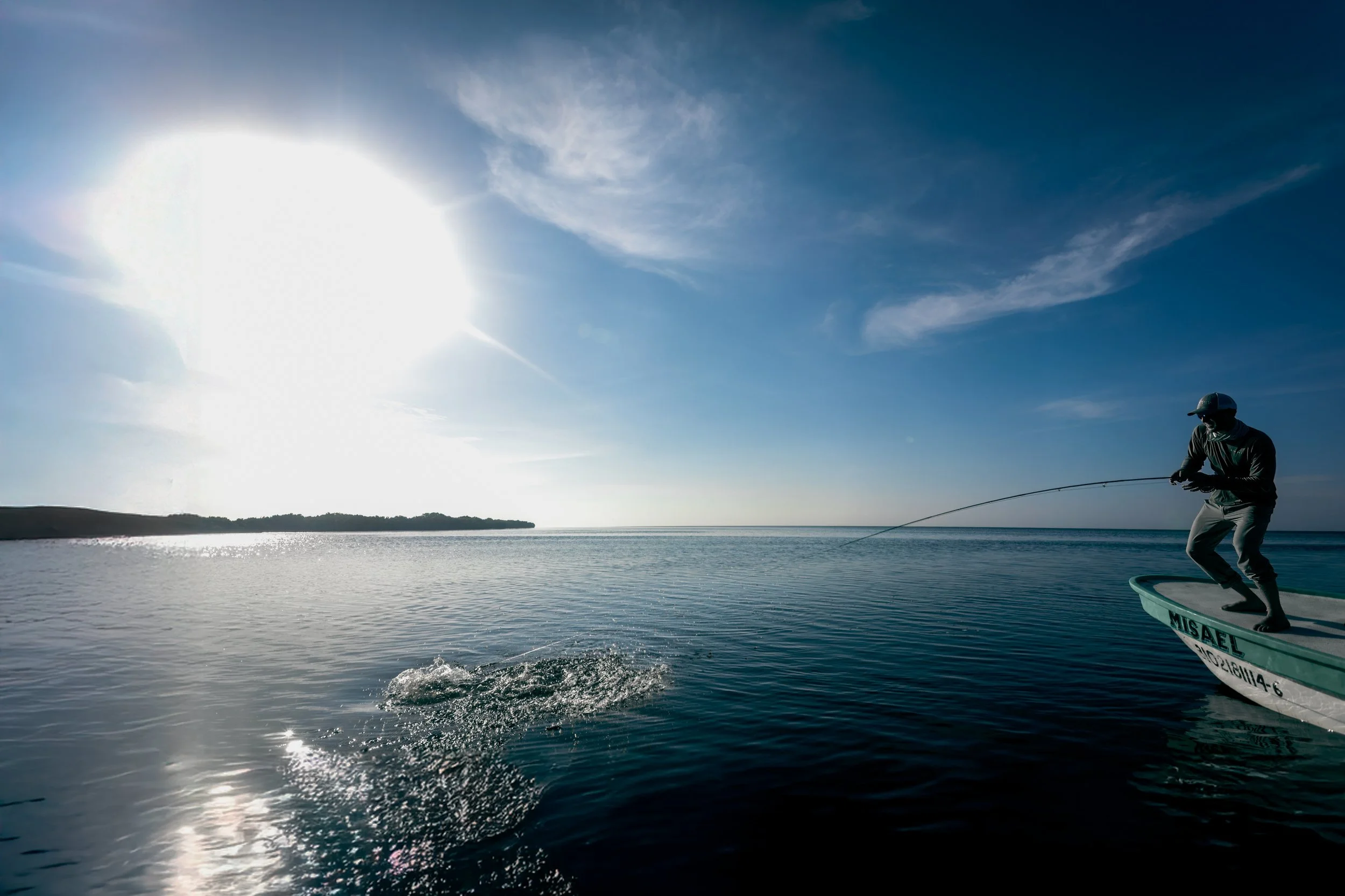 A person fishing on a boat in a large body of water during sunset or sunrise, with the sun low on the horizon and a clear sky with some clouds.