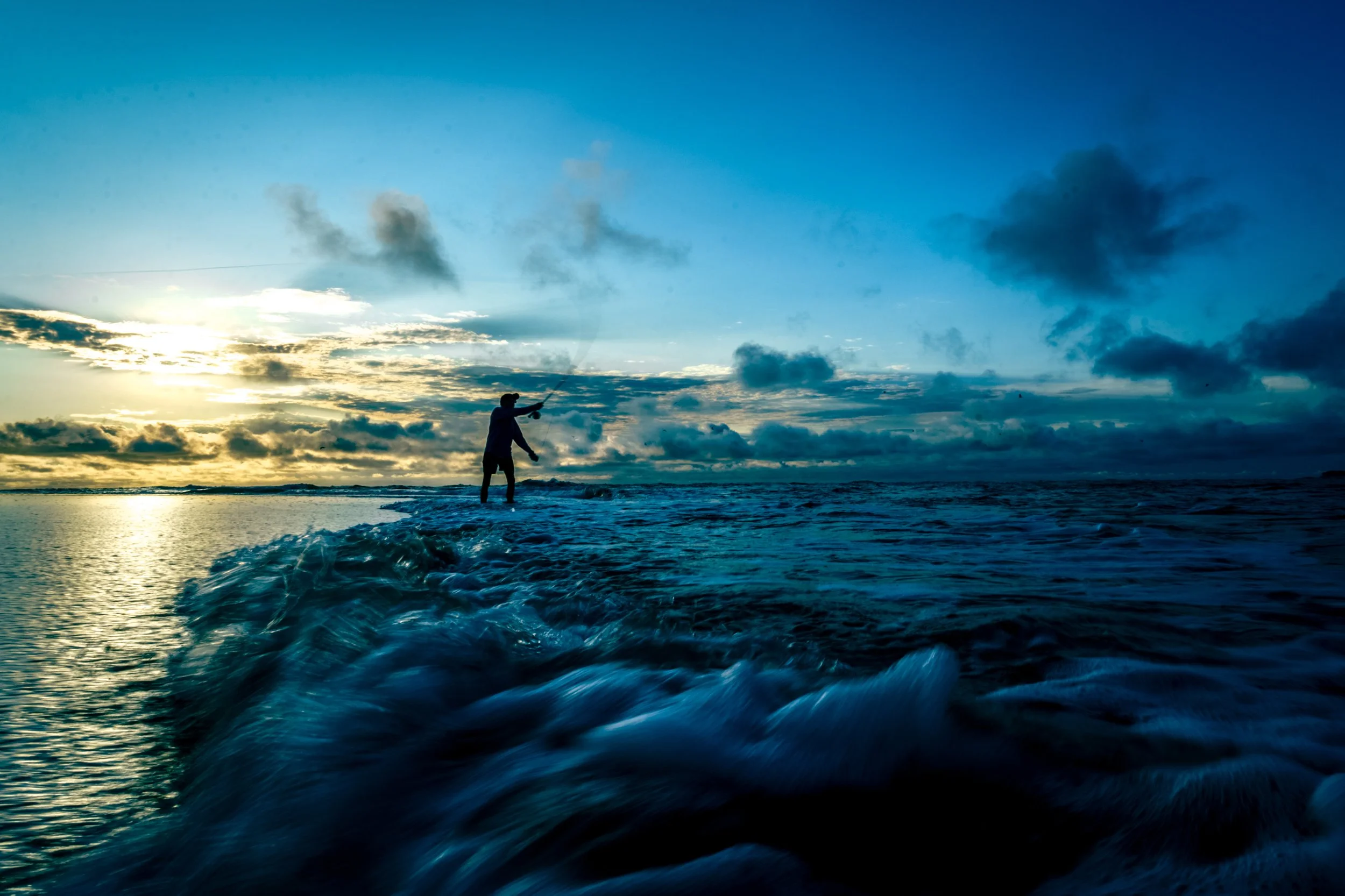 A person standing on a boat or surfboard in the ocean, holding a fishing rod, with the sunset or sunrise sky and clouds in the background.