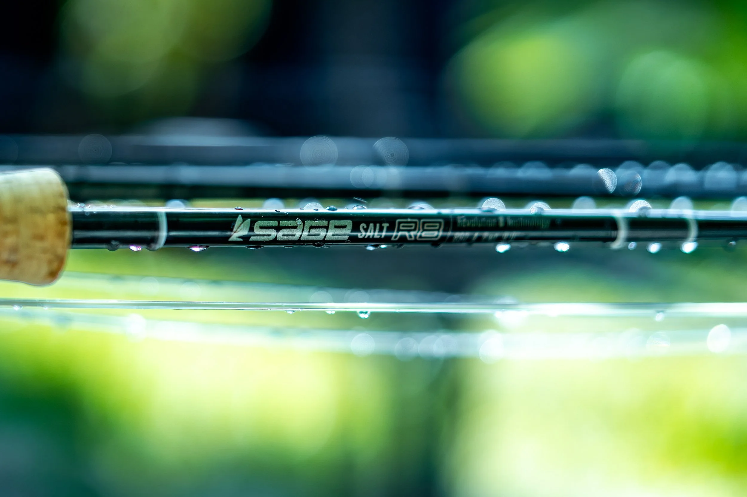 Close-up of a black fishing rod with a cork handle, with water droplets on the rod, set against a blurred green outdoor background.