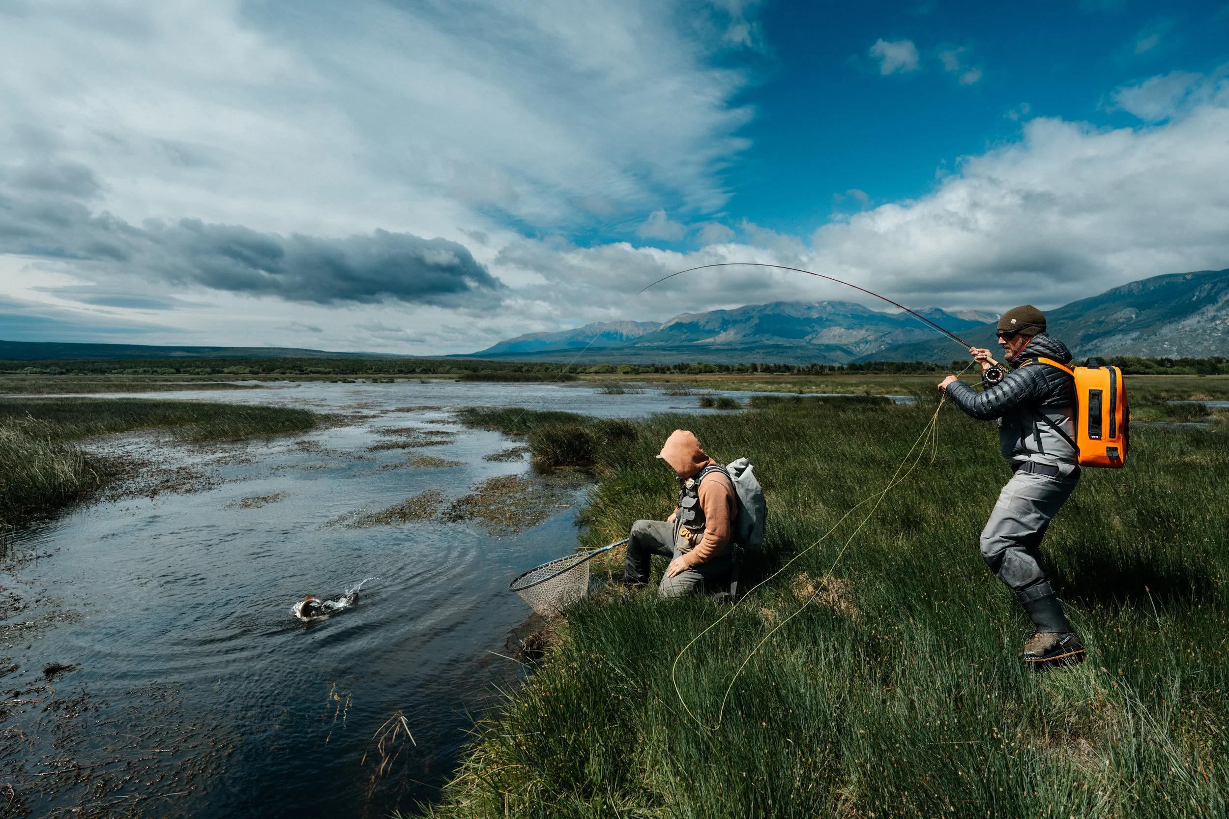 A guide and angler landing a trout in Patagonia with mountains in the background during a cloudy day.