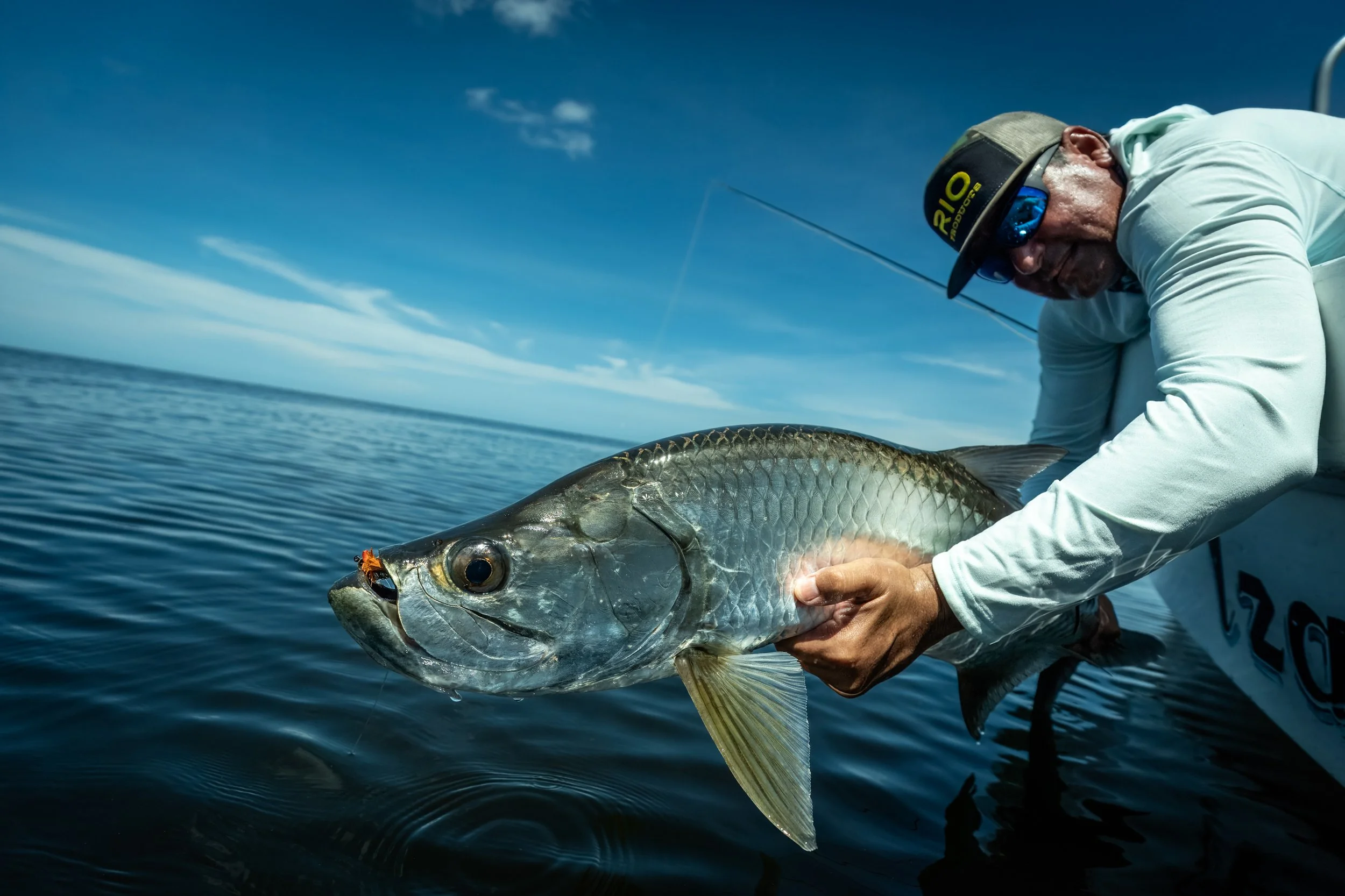 A person wearing a cap, sunglasses, and a long-sleeve shirt is holding a large tarpon over water on a boat, with a backdrop of blue sky and distant clouds.