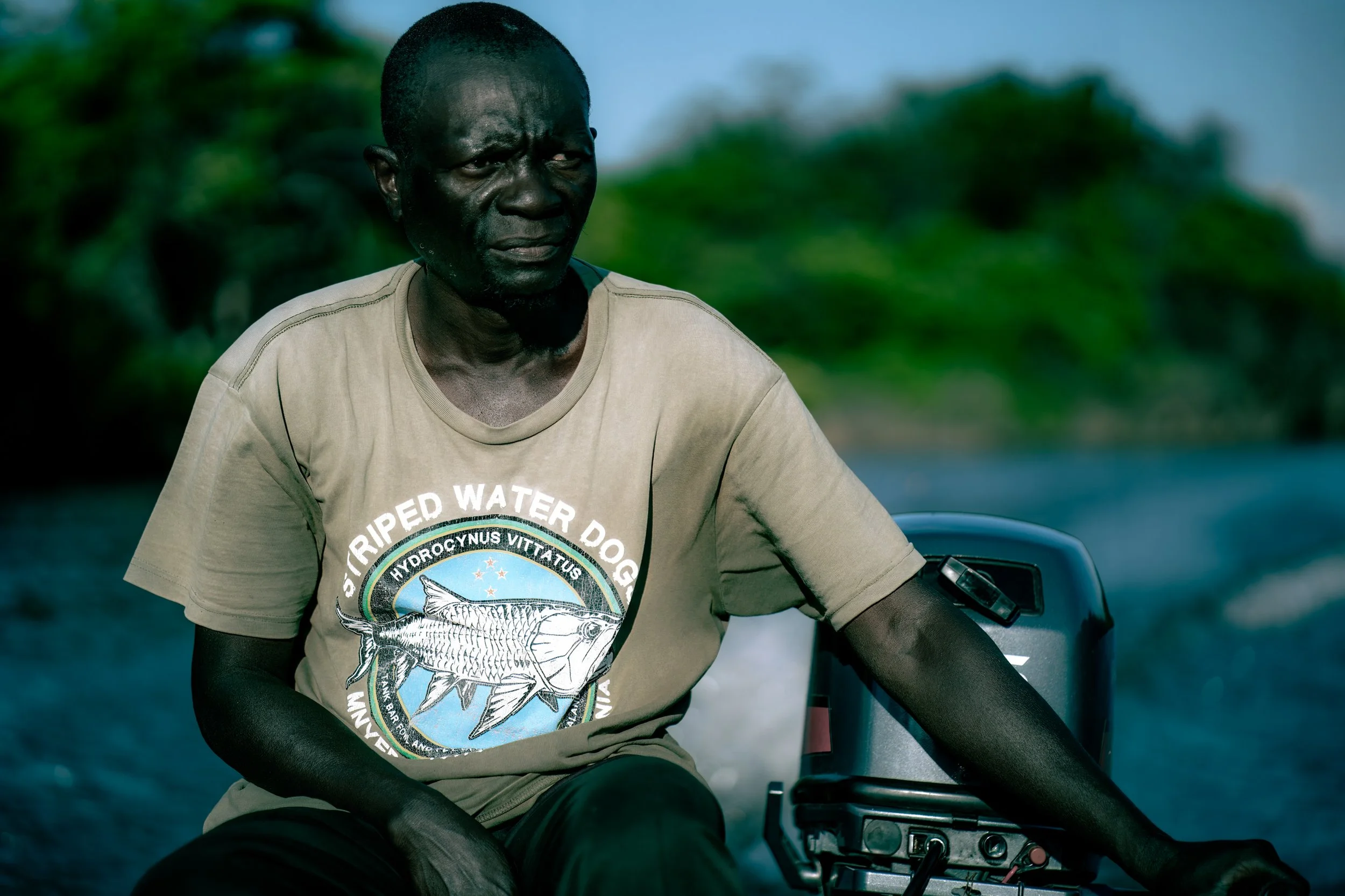 A man with dark skin sitting in a boat on a body of water, wearing a beige T-shirt with a fish graphic and text, with a scenic background of greenery and blue sky.