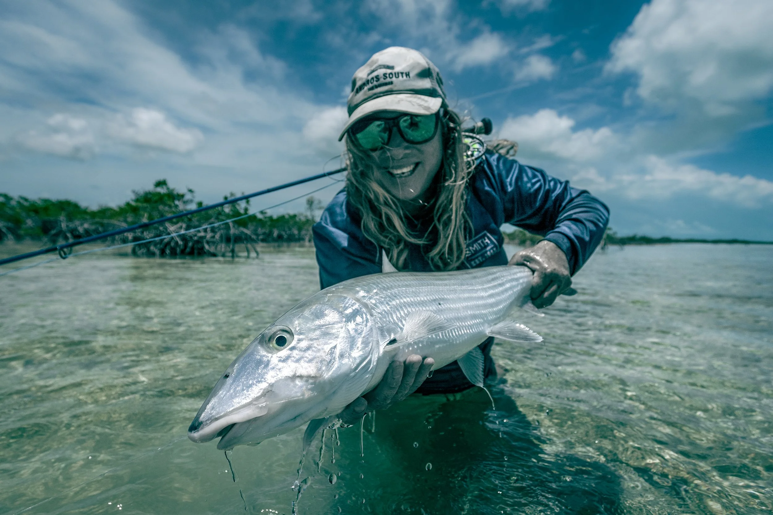 Person wearing sunglasses, a cap, and a blue jacket holding a fish in shallow water, with a partly cloudy sky and trees in the background.