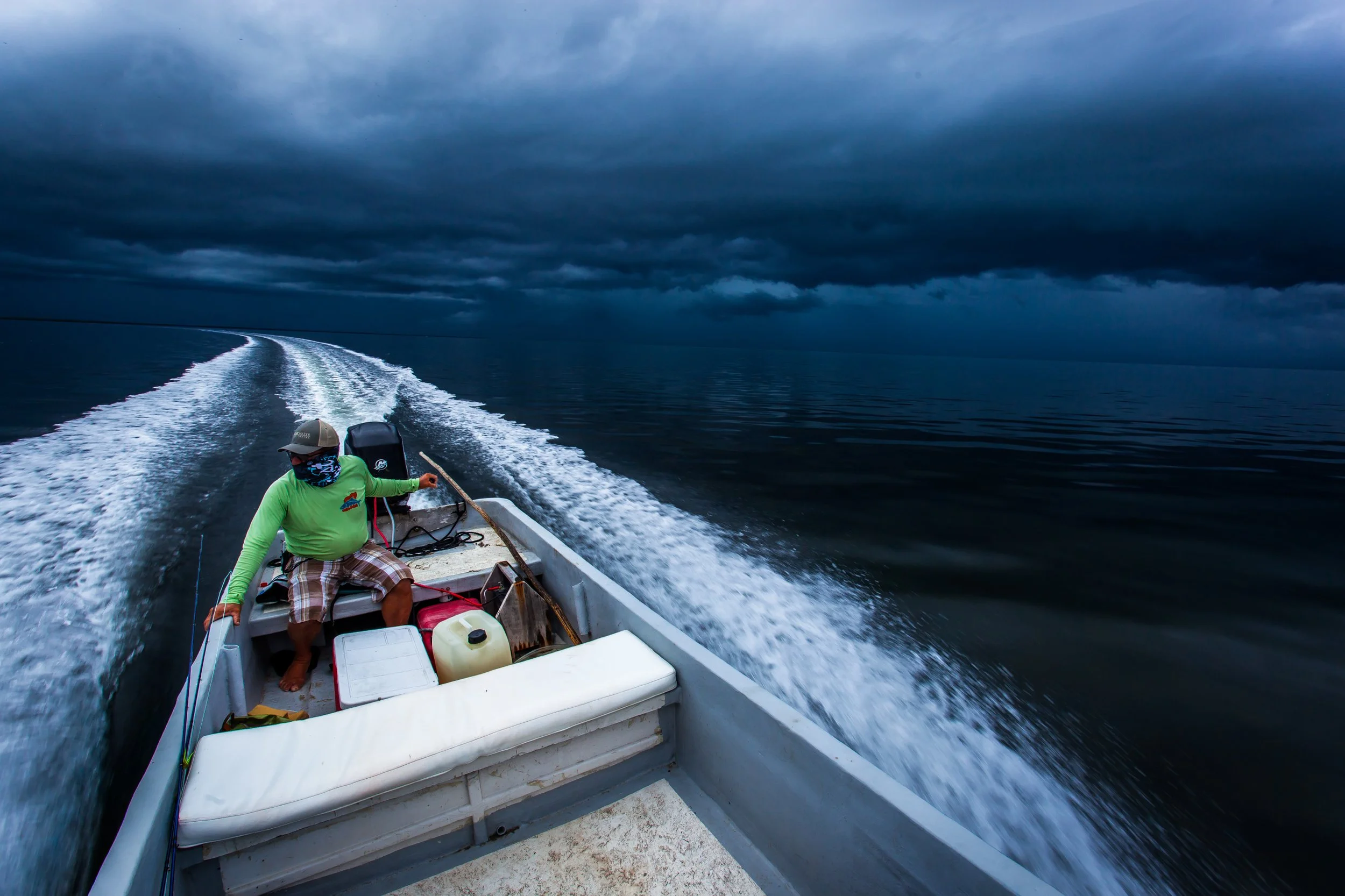 A guide sitting at the front of a small boat on dark waters during cloudy weather, with storm clouds overhead and a wake behind the boat.