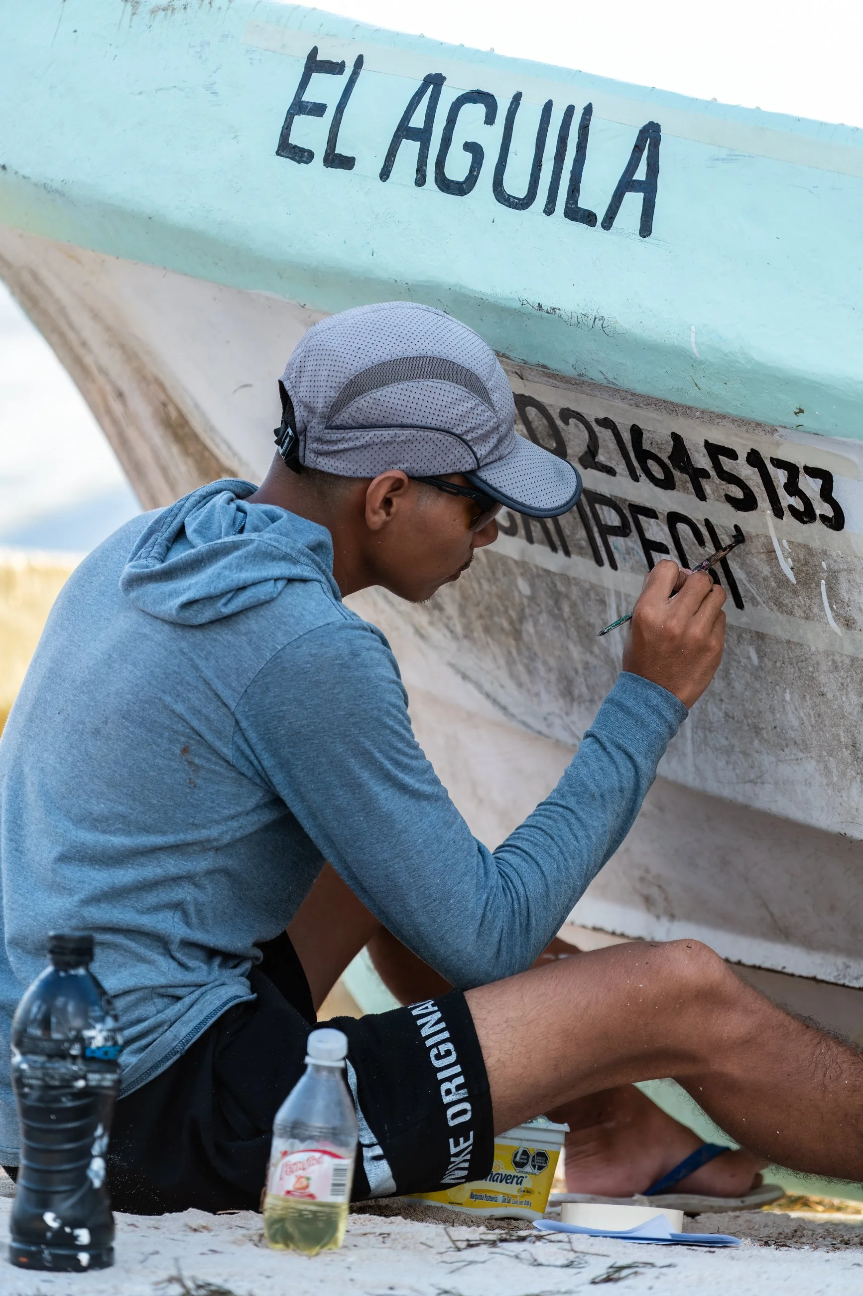 A man in athletic clothing and a gray cap is sitting on the ground and painting a large sign that says 'EL AGUILA' on a boat or large surfboard, with two bottles of water nearby. in Mexico.