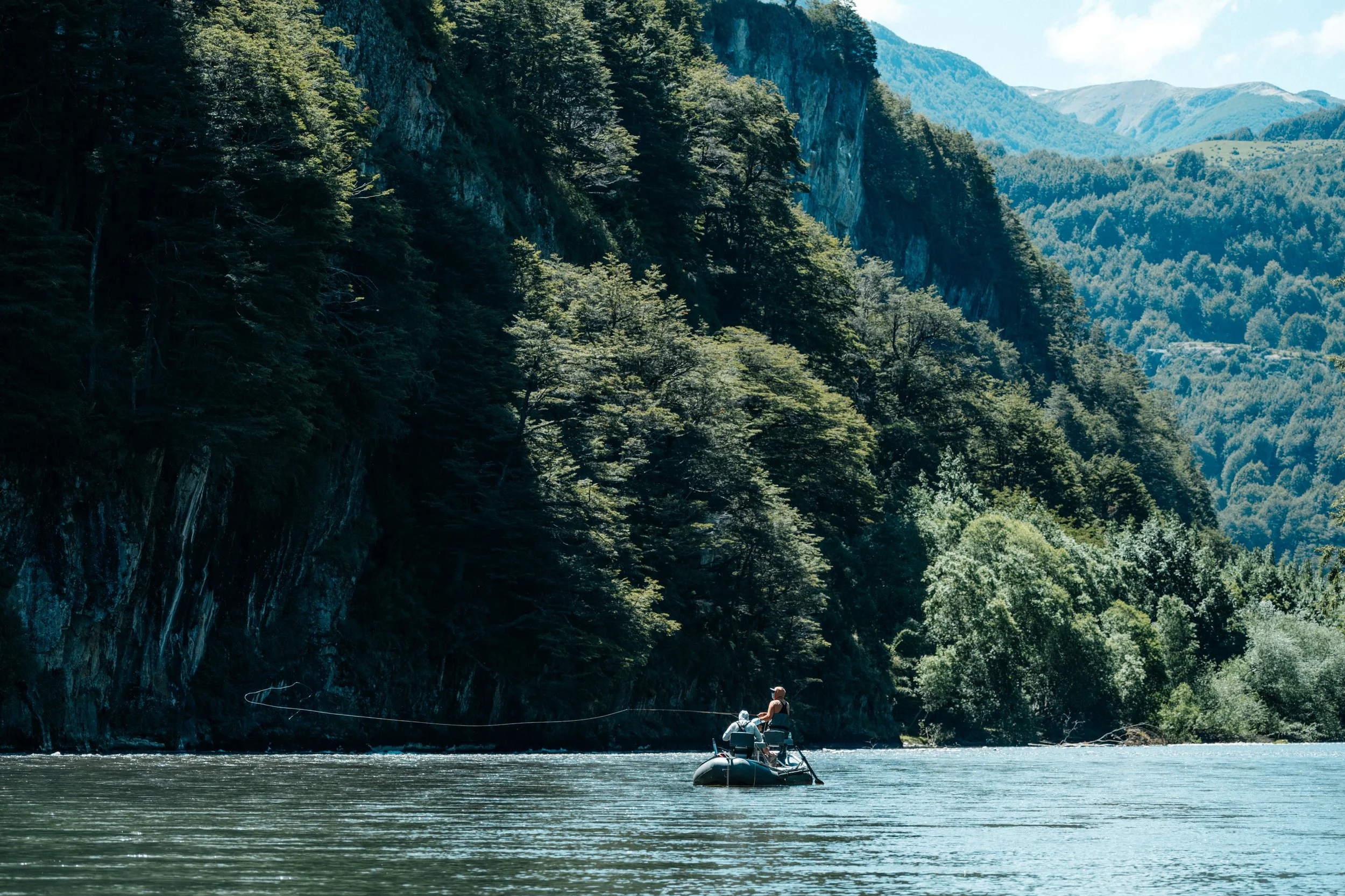 People fly fishing from a boat on a river in Chile surrounded by tall, green forested mountains.