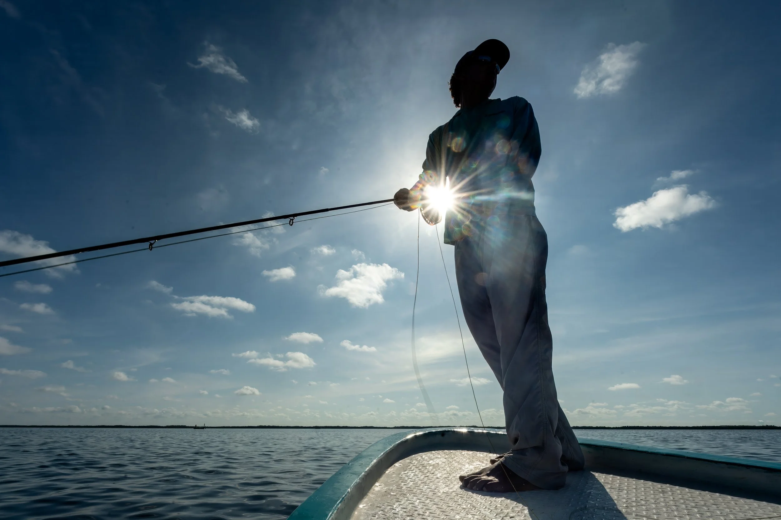 Person fishing on a boat in a large body of water with a sky filled with clouds, backlit by the sun, casting a glow around the person.