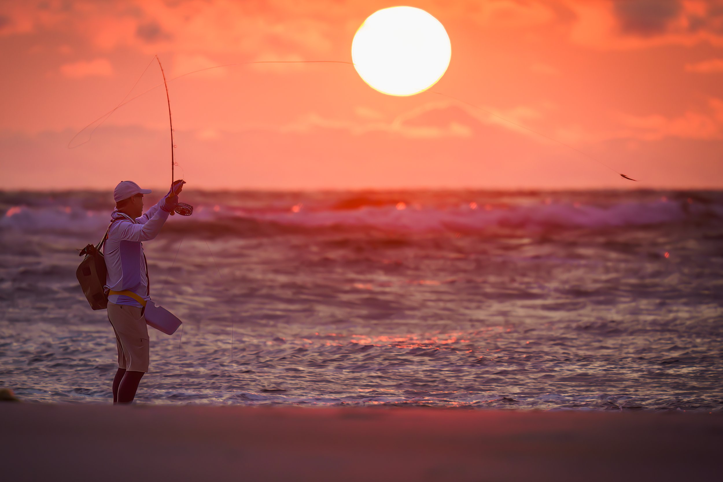 Person fly fishing in the ocean during sunset with a large sun in the sky.