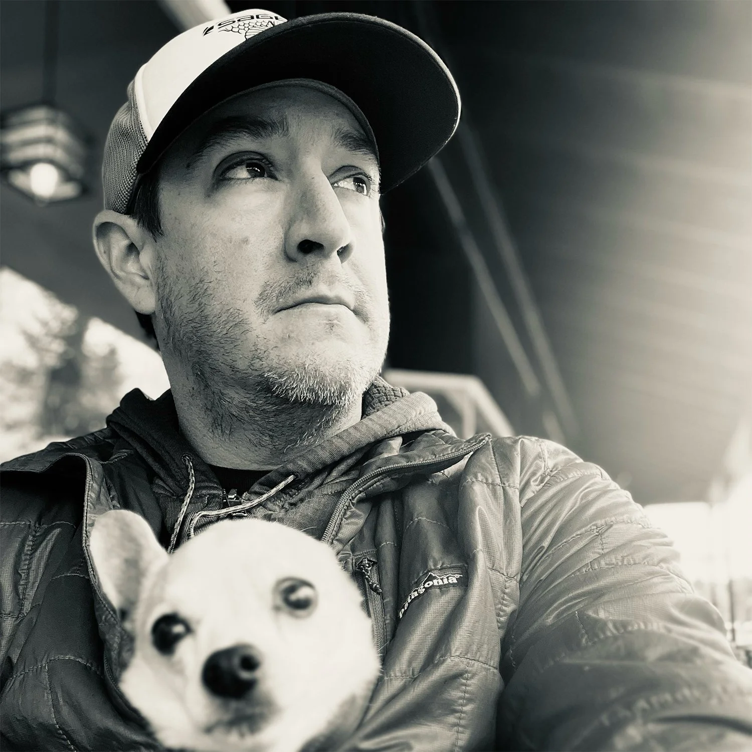 A black and white photo of a man wearing a baseball cap and puffy jacket, holding a small dog with white and black fur, inside a vehicle.