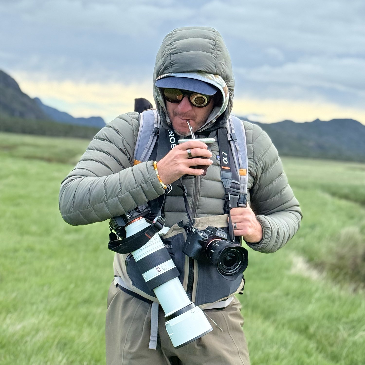 A man dressed in outdoor gear, including a hoodie, sunglasses, and a cap, holding a camera with a large telephoto lens around his neck, and sipping from a small cup in a green open field with mountains and cloudy sky in the background.