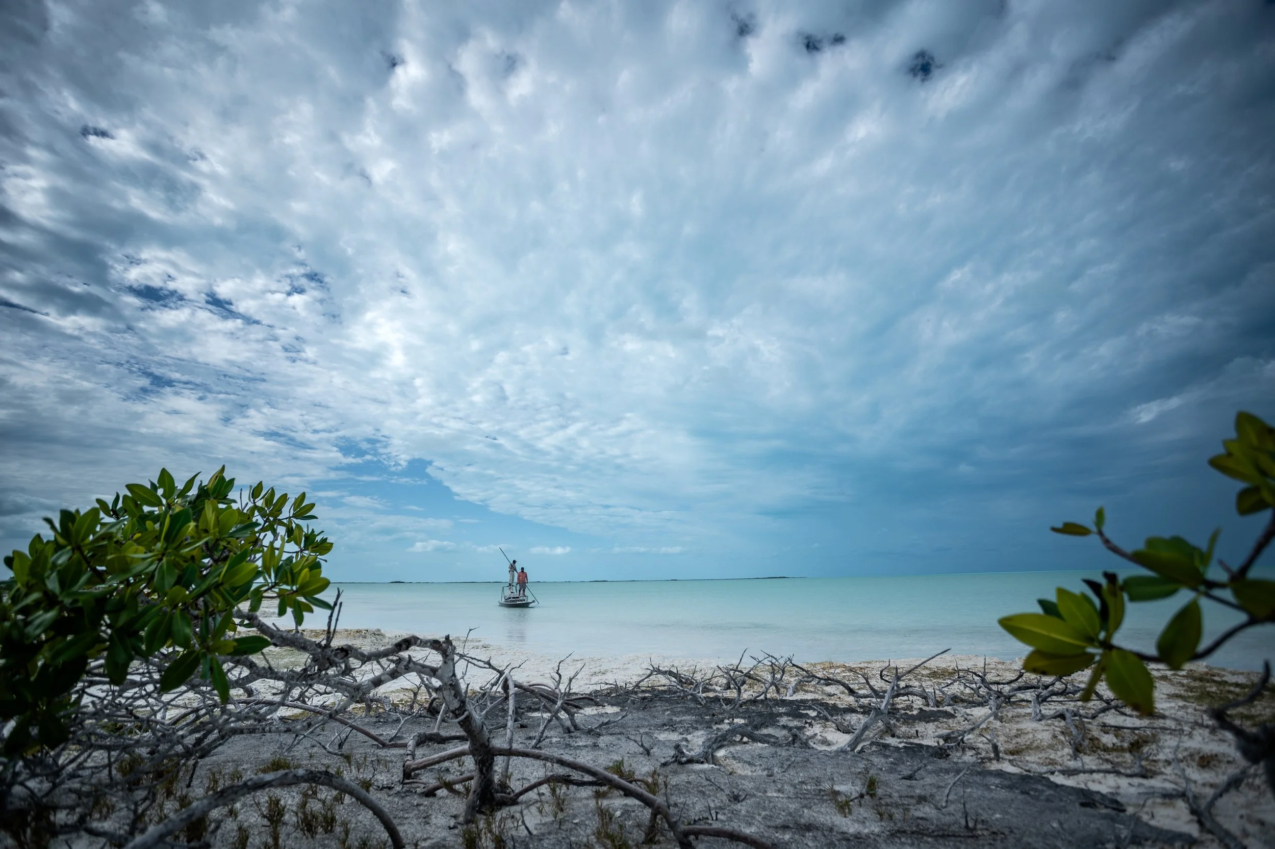 View of a bonefish flat in the Bahamas with clear turquoise water, some clouds in the sky, and a person on a boat in the distance, surrounded by greenery.