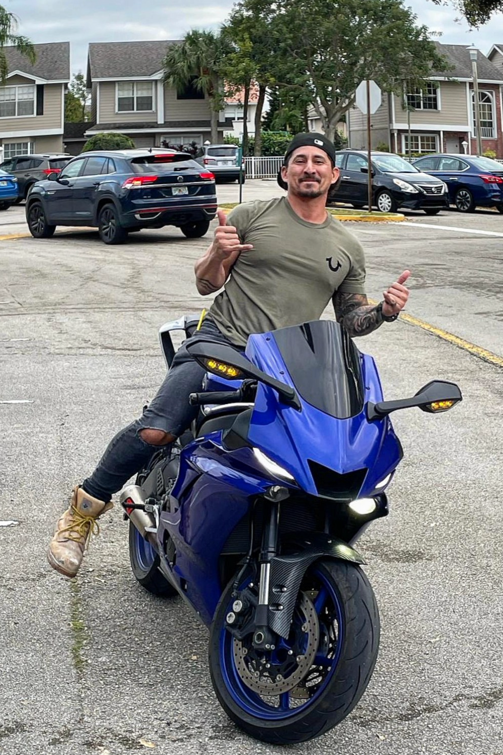 A man wearing a black baseball cap, an olive green T-shirt, and ripped black jeans is sitting on a blue motorcycle in a parking lot, smiling and making a hand gesture.