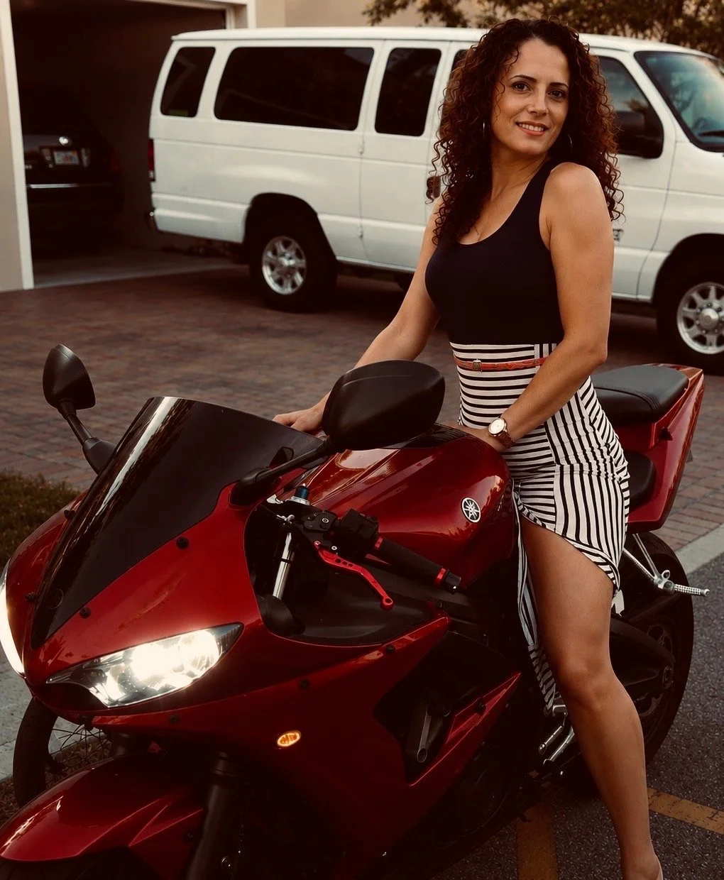 A woman with curly hair and a striped skirt sitting on a red sport motorcycle in a parking lot, with a white van and other vehicles in the background.