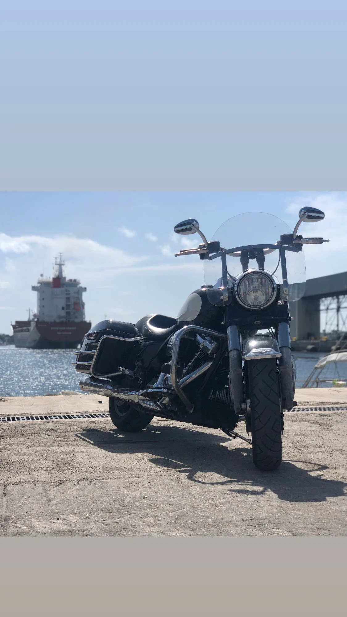 Black motorcycle parked near water with a cargo ship in the background under a partly cloudy sky.