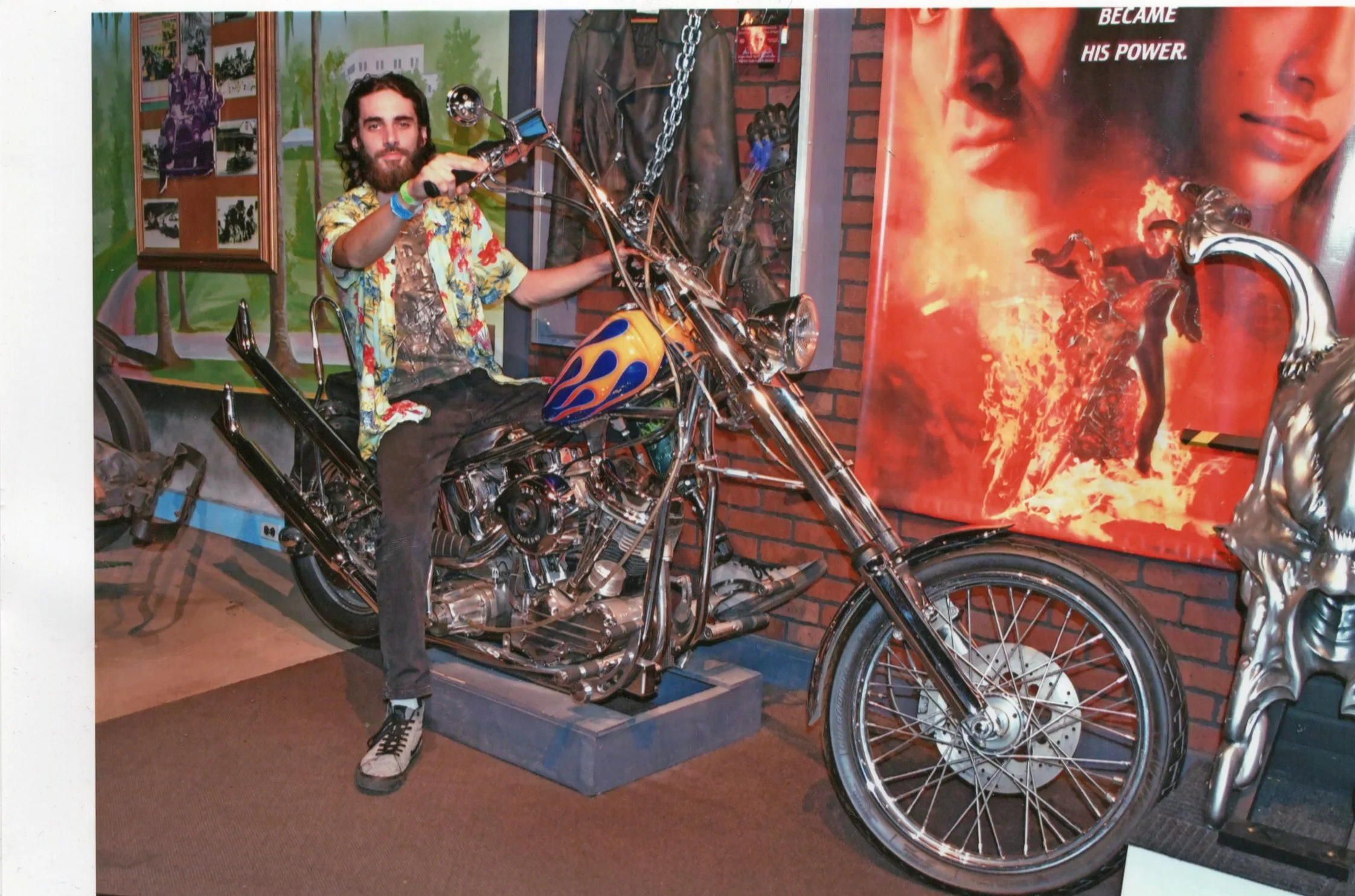 A man with long hair and a beard is sitting on a motorcycle inside a room. He is wearing a colorful floral shirt, black pants, and white sneakers. The motorcycle has a shiny chrome finish with flames painted on the gas tank. Behind him, there is a brick wall with a large poster of a woman's face and some chains hanging. To the left, there is a green mural with trees and homes. The room appears to be a display or museum with various items and posters.