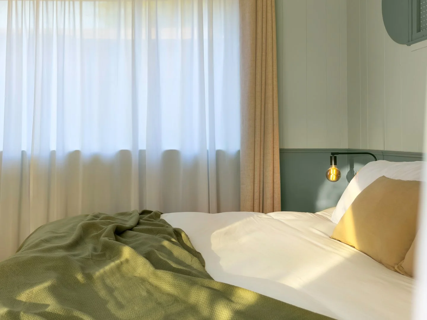 A bed with white sheets, yellow pillows, and a green blanket in a bedroom with large white curtains covering the window. The wall behind the bed is painted light green with a dark green trim.