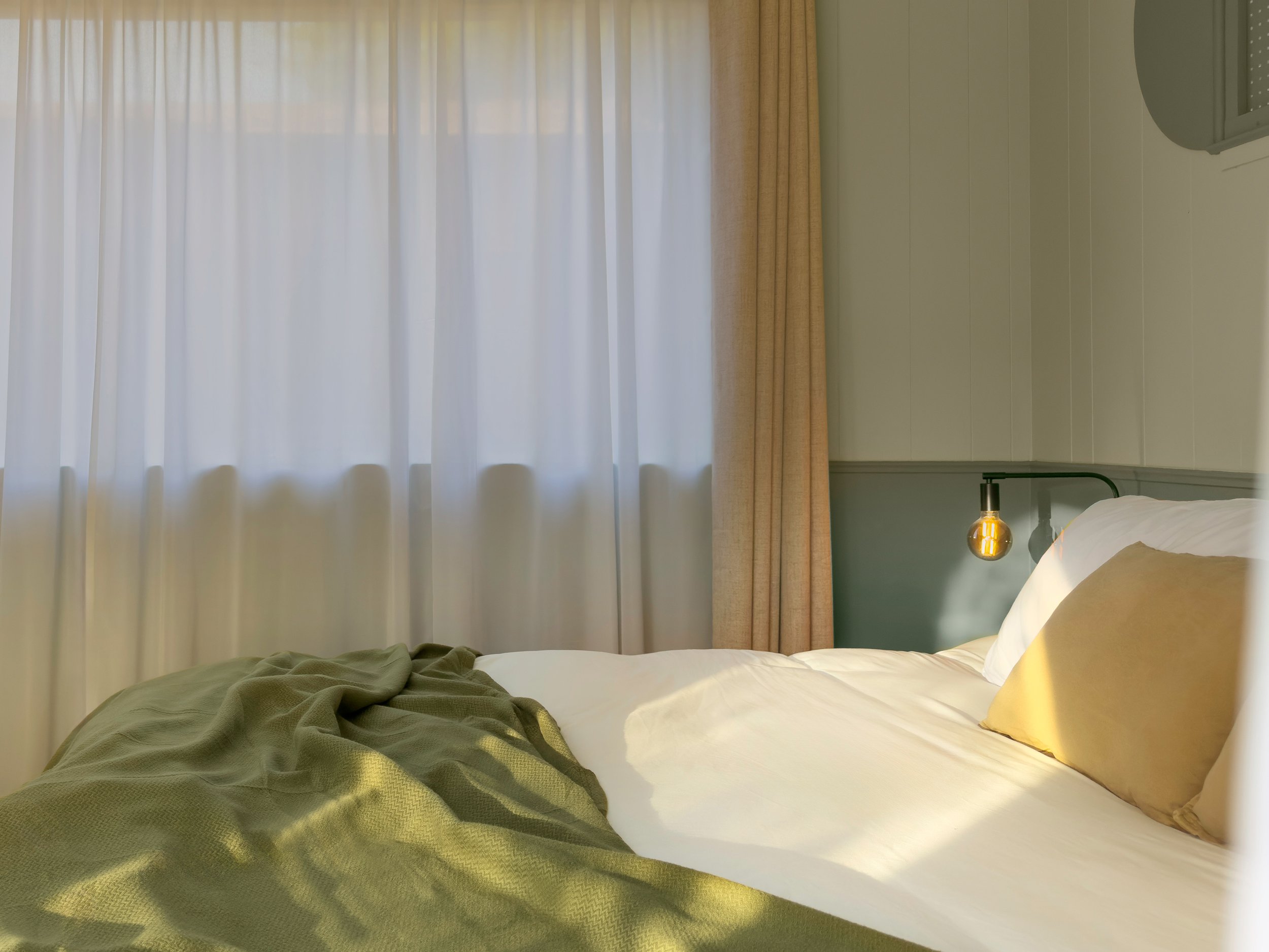 A neatly made bed in a hotel room with white sheets, a beige pillow, and a green blanket, against a background of white curtains and a wall-mounted lamp.