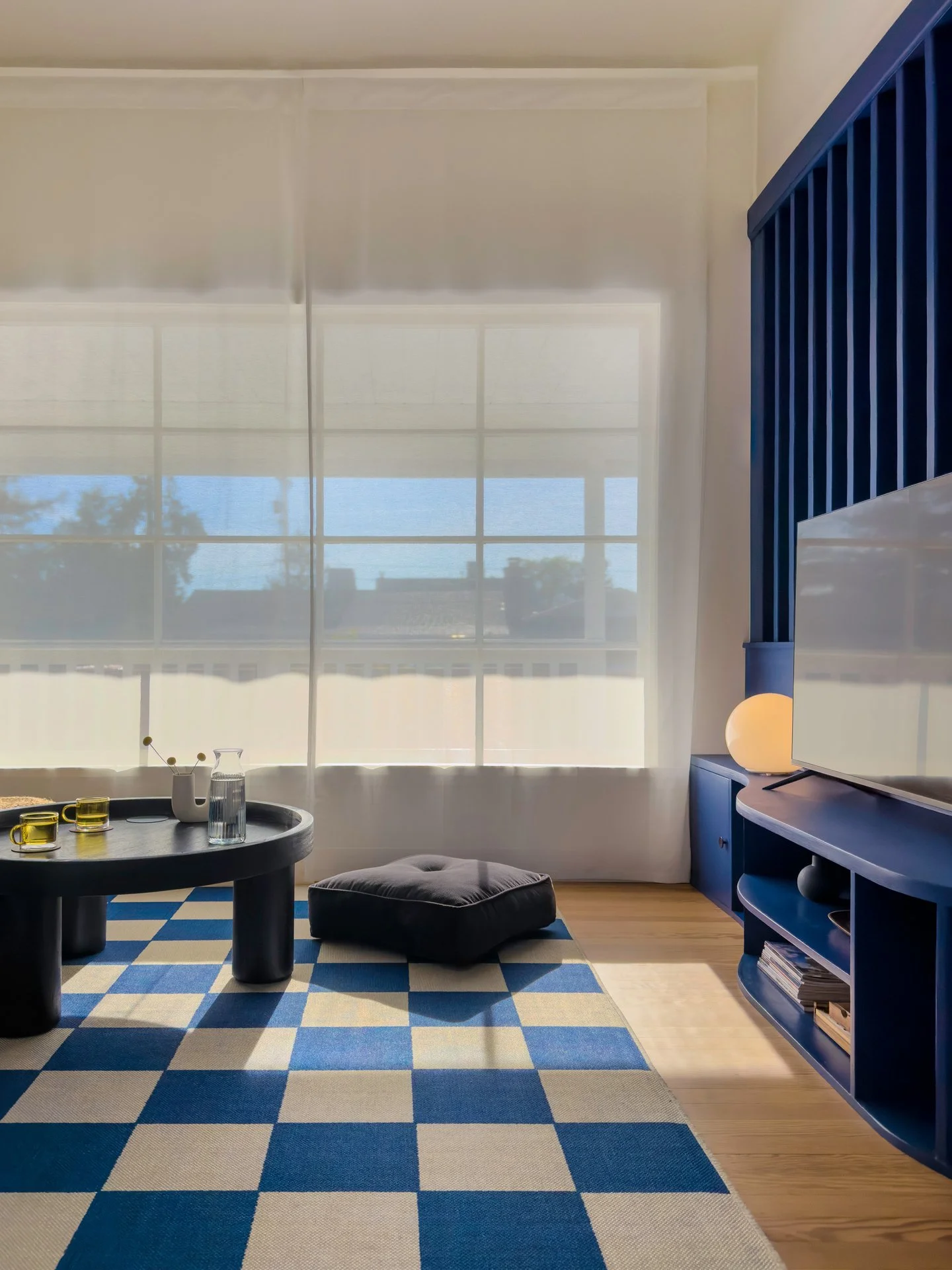 Living room with a checkered blue and beige rug, a round coffee table with yellow glasses and a pitcher, a gray cushion on the floor, a window with white blinds, a blue entertainment center with a TV and decorative items, and a small lamp.