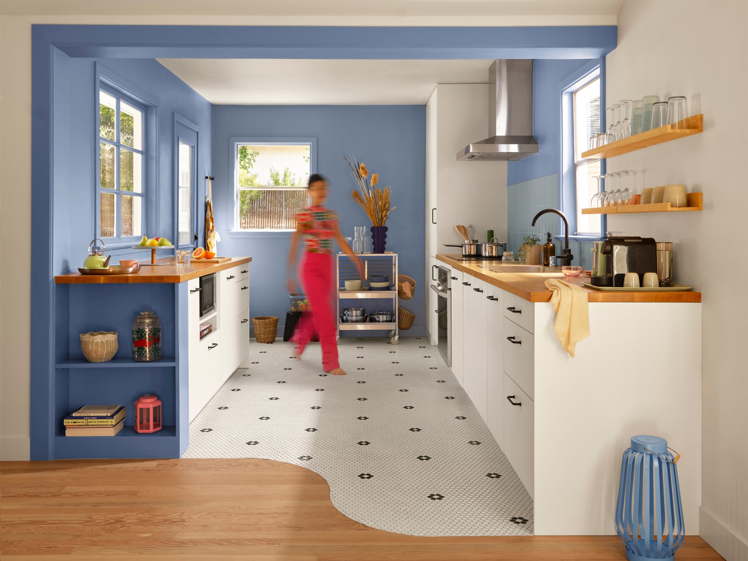 A kitchen with white cabinets and blue walls. There are large windows, a blue shelf with books and decor, a wooden countertop with fruit and a teapot, and a person in a floral shirt and pink pants walking through the space.