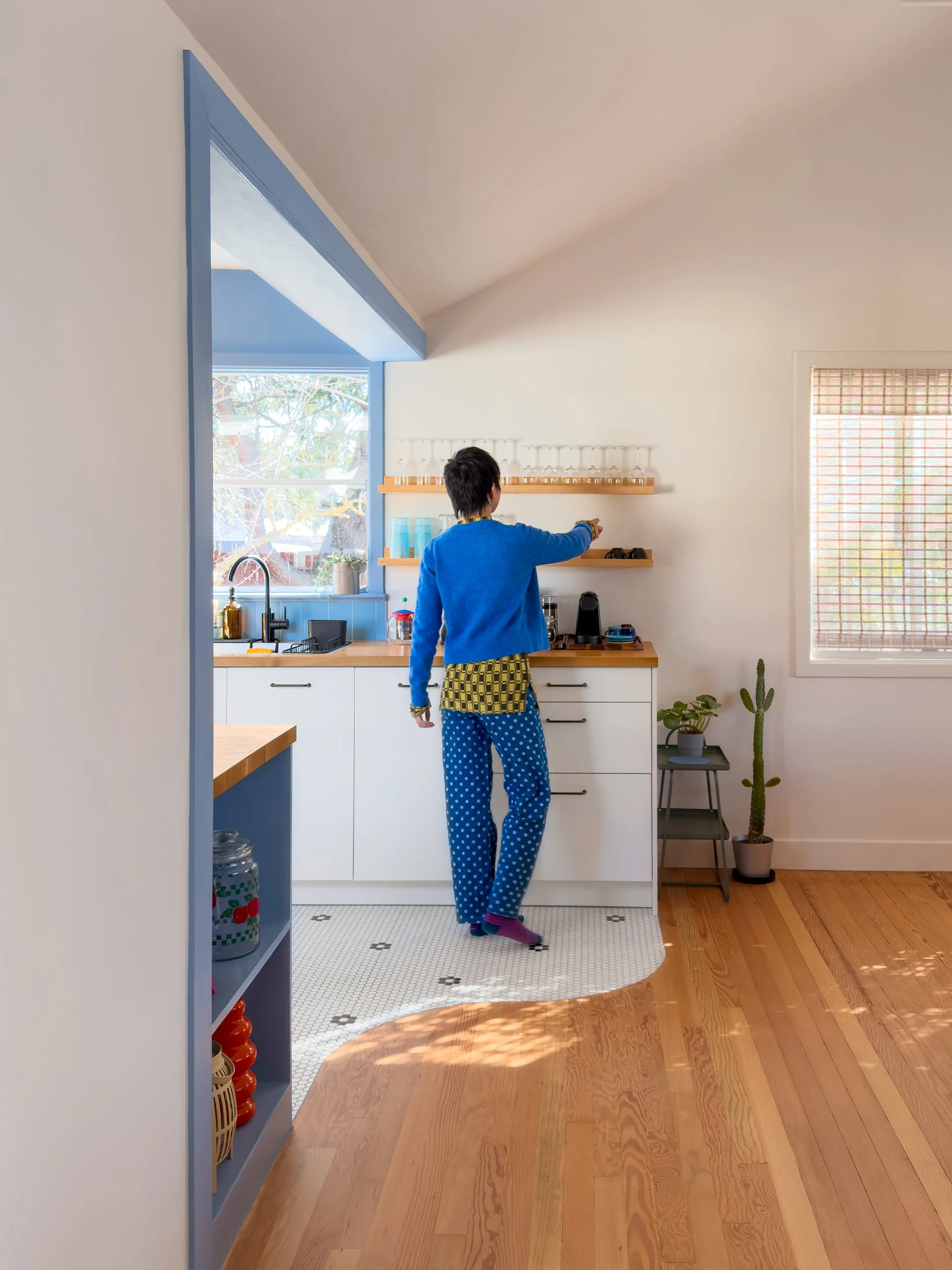 A person with short dark hair wearing a blue sweatshirt, yellow checkered apron, and blue polka dot pajama pants is standing in a small kitchen, reaching towards a shelf. The kitchen has white cabinets, a large window with a view of trees, and minimal decor.
