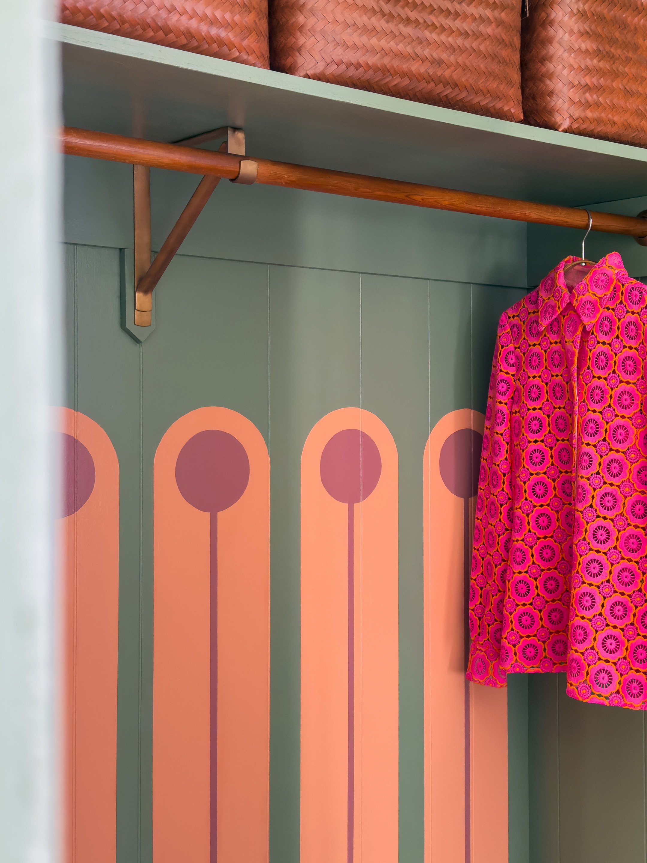 Inside a closet, a pink floral dress is hanging on a wooden rod. The closet has a geometric, colorful wall design with circles in shades of pink, orange, and purple.