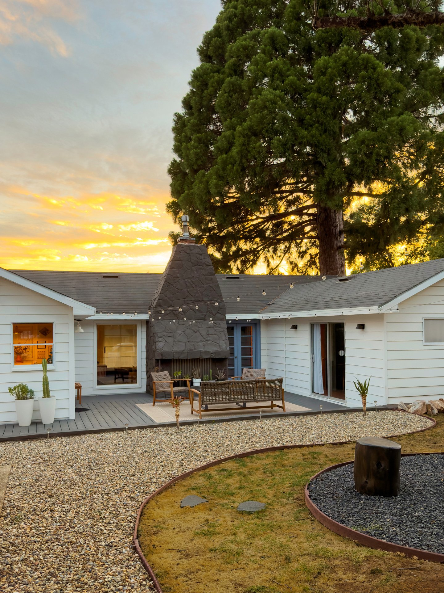 A backyard patio with outdoor seating and a stone fireplace under a large tree at sunset.