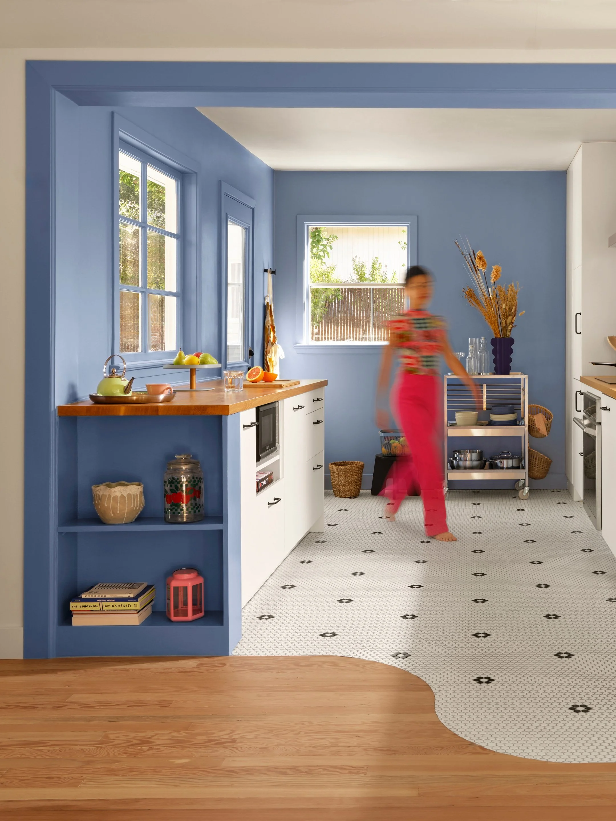 A brightly lit kitchen with blue walls and white cabinets. A woman in a colorful top and pink pants is walking across the mosaic tile floor, which transitions from hardwood to a black and white pattern. There are windows showing greenery outside and a blue vase with dried flowers on a shelf.