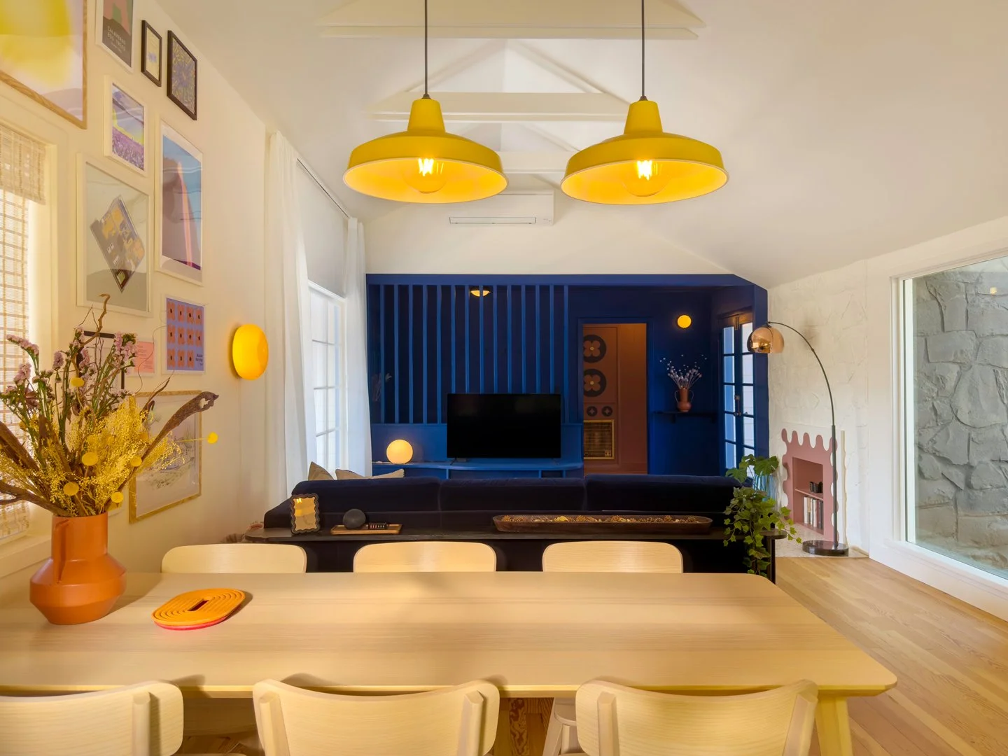 A dining room with a kitchen in the background. There is a long white table with white chairs, a large yellow vase with dried flowers, and a stack of colorful coasters. The room has framed artwork on the wall, yellow pendant lights hanging from the ceiling, and a television on a blue accent wall.