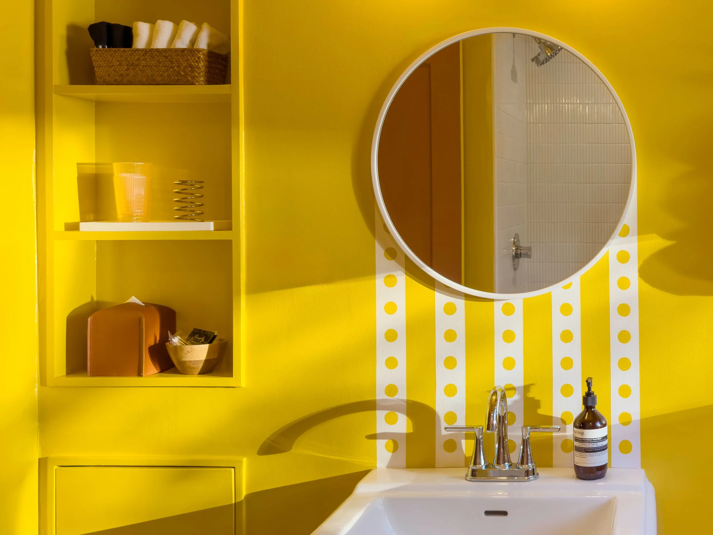 A yellow bathroom with a white sink, round mirror, and yellow shelves. The shelves contain black and white towels, yellow and orange containers, and a metallic spring-like object. A soap dispenser is on the sink.