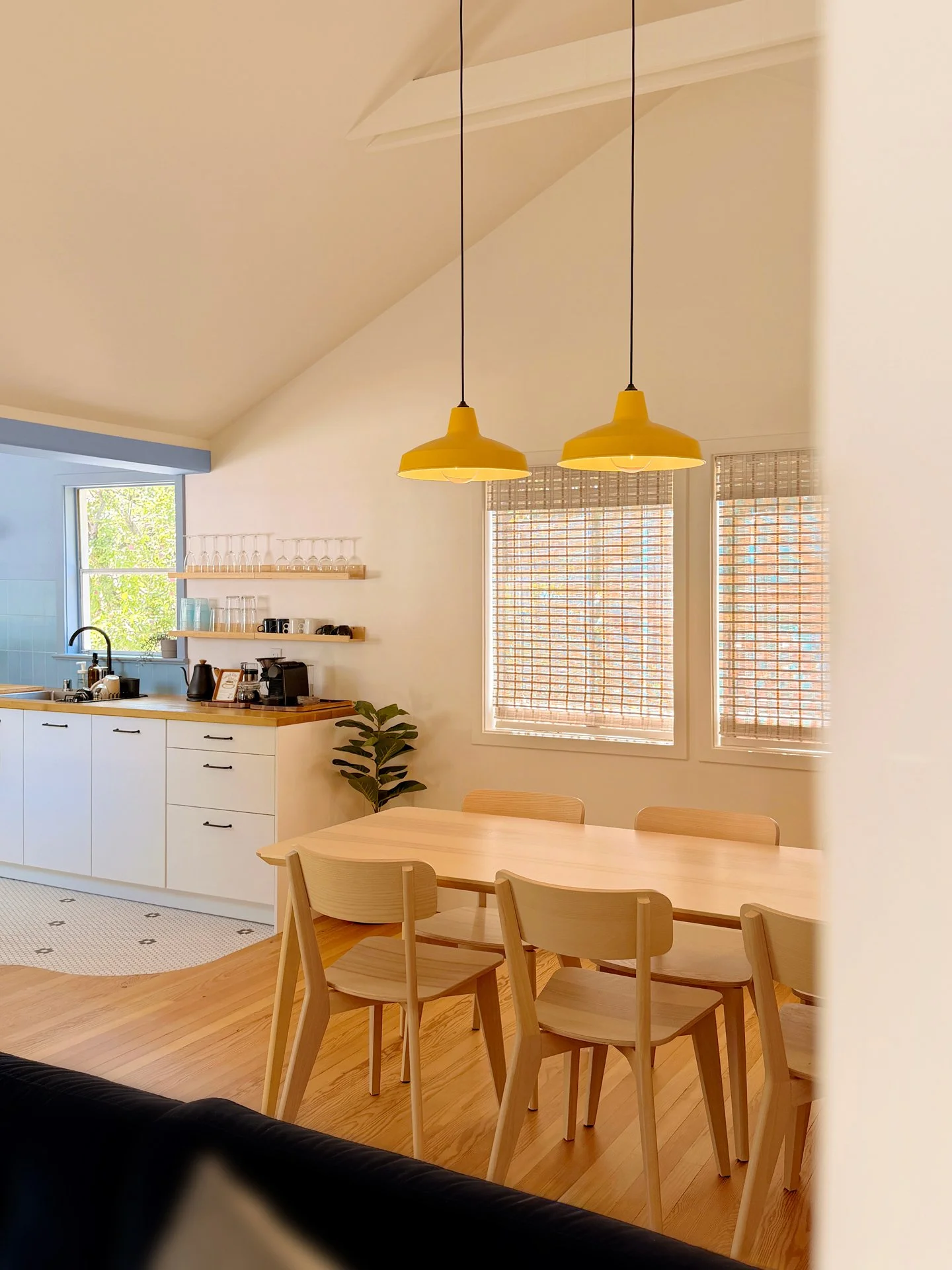 Interior of a modern kitchen and dining area with natural light, wooden furniture, hanging yellow pendant lights, and bamboo window shades.
