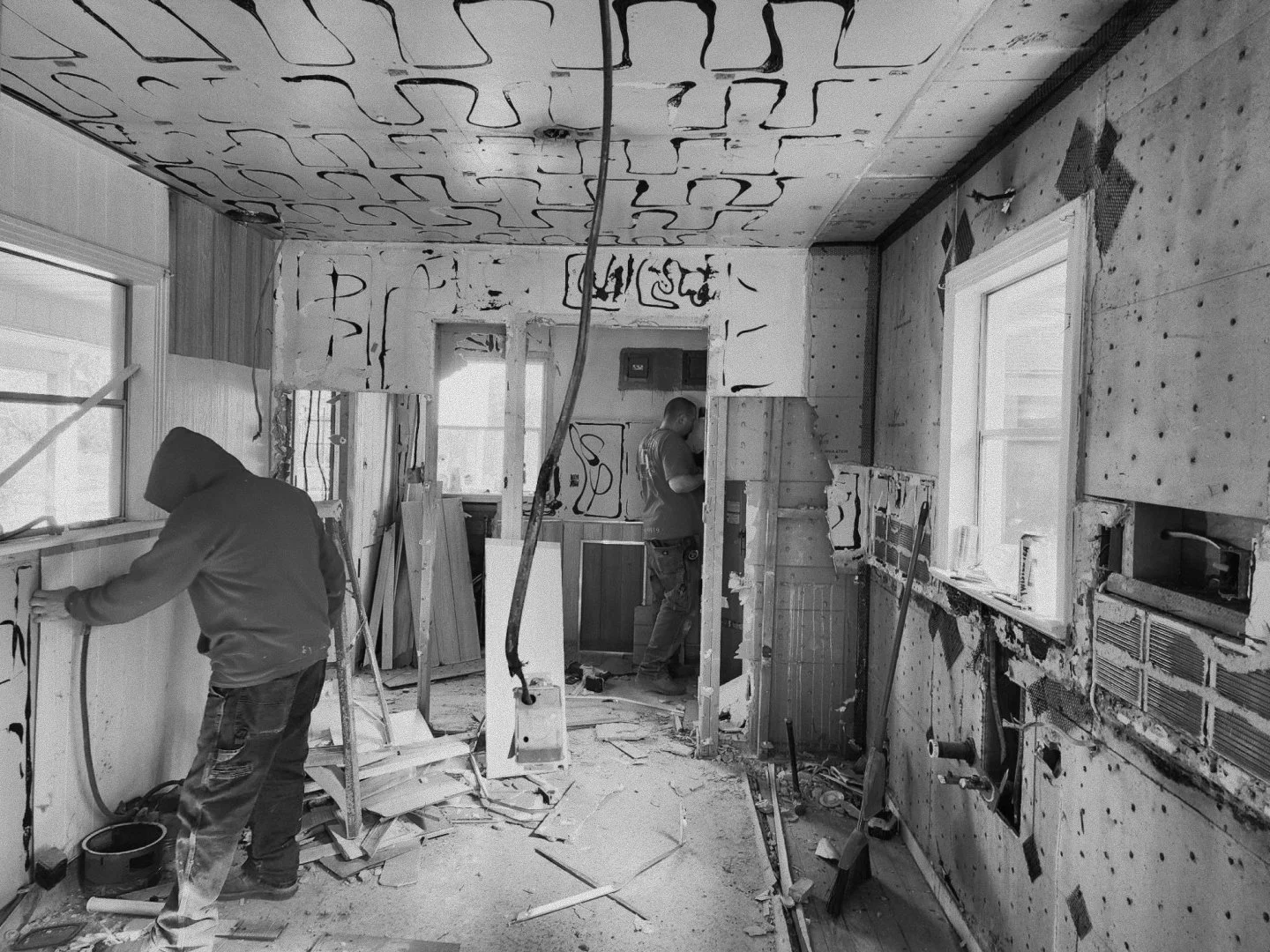 Two workers renovating a room with drywall and windows, construction tools and debris scattered on the floor.